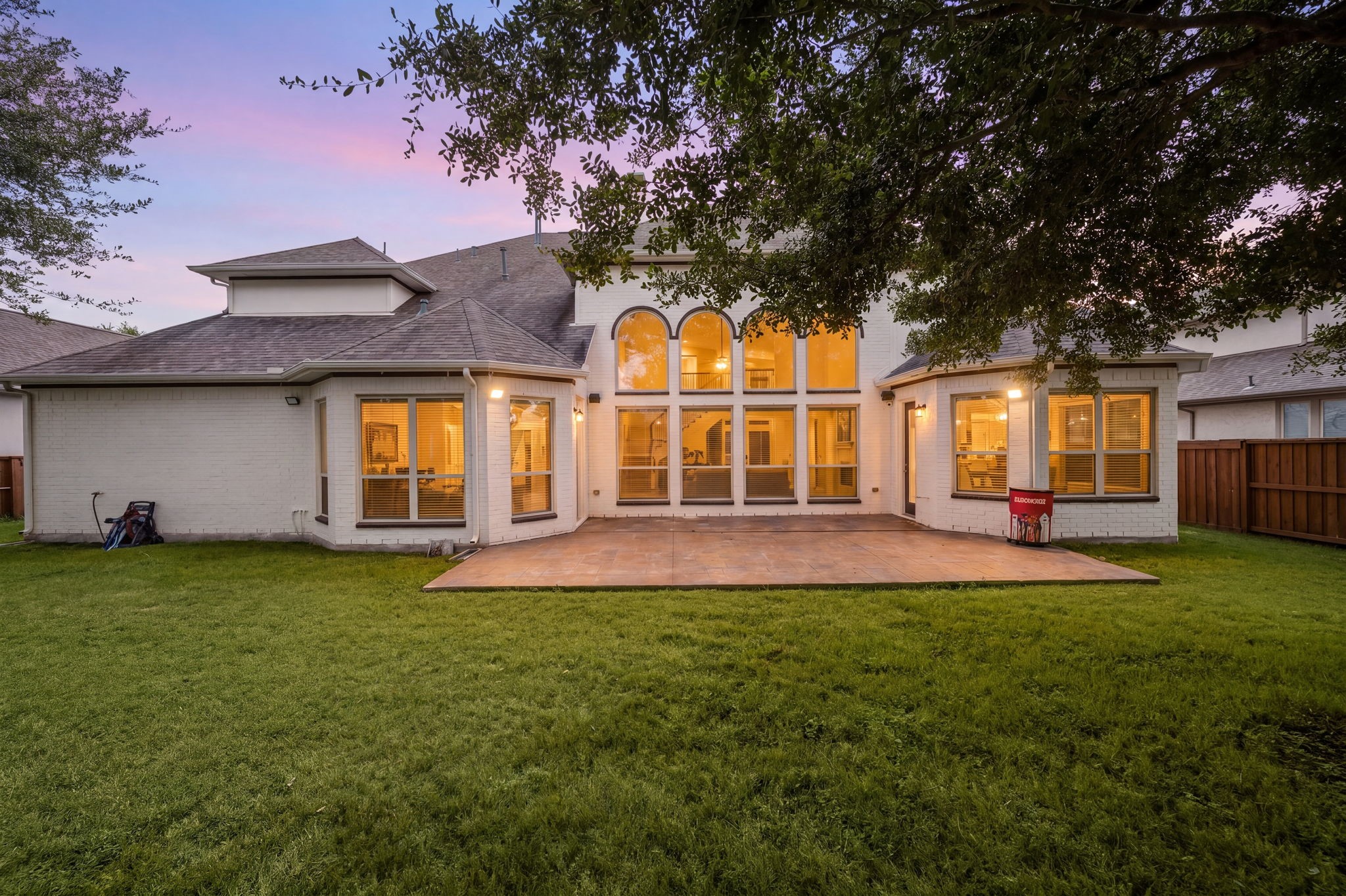 At dusk, the home's dramatic arched two-story windows glow from within, overlooking a covered patio and expansive backyard framed by mature trees.
