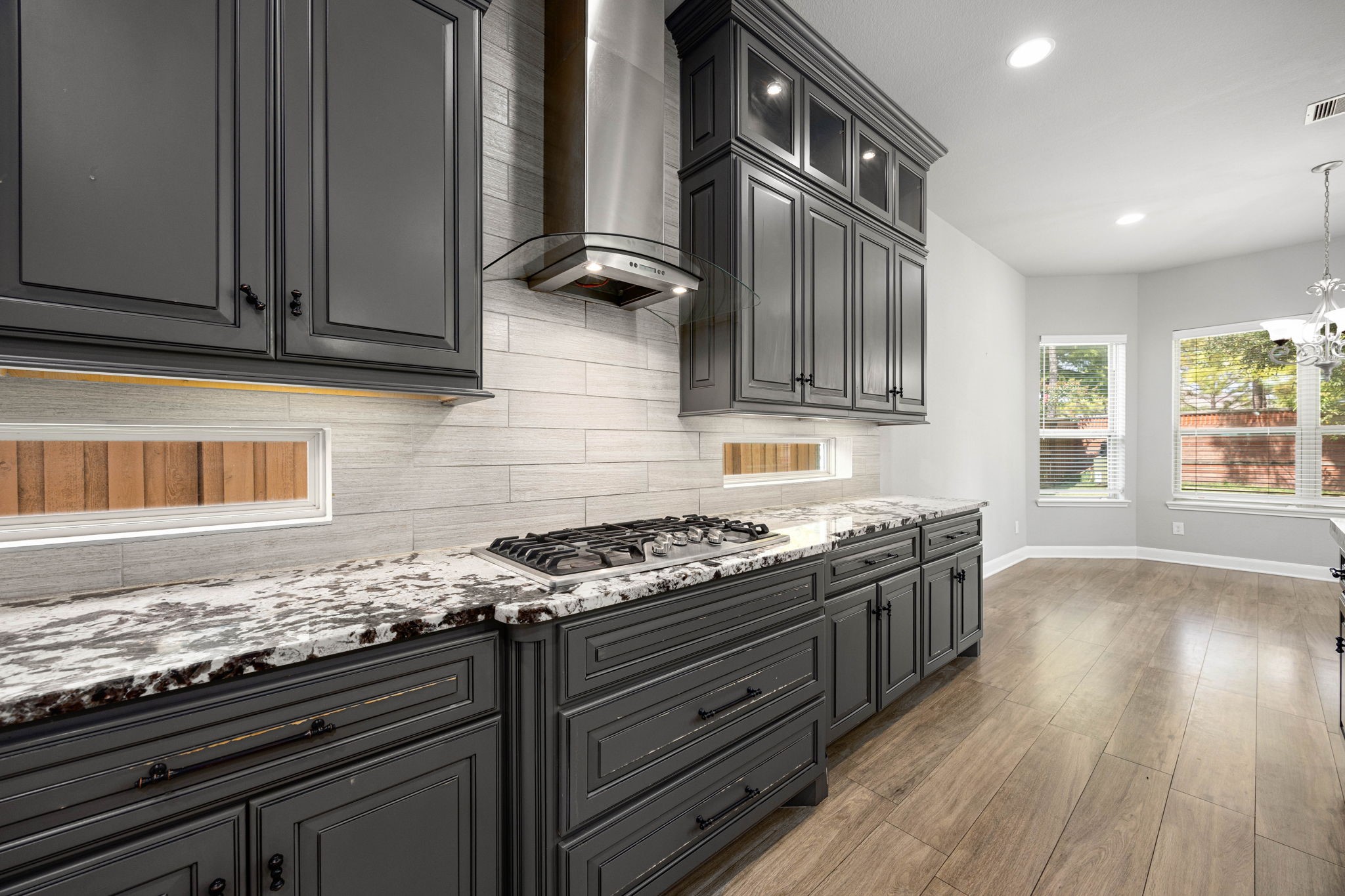 A gas cooktop beneath a sleek glass-and-steel range hood anchors this section of the kitchen, framed by marble counters and a tile backsplash that extends throughout the space.