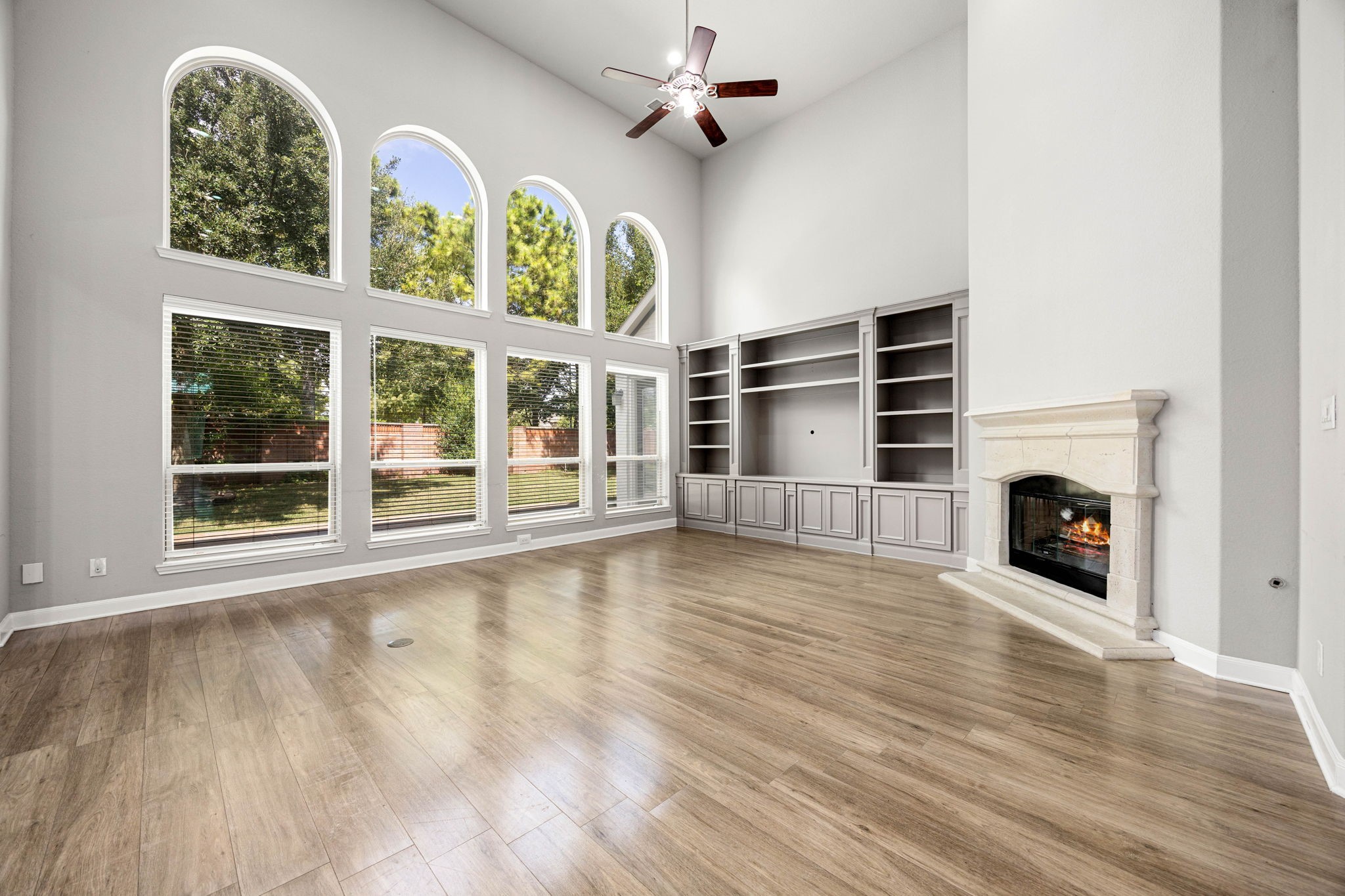 Soaring ceilings and a wall of arched windows fill this open living area with natural light, anchored by a stone fireplace and custom built-in shelving, a bright, modern centerpiece for the home.