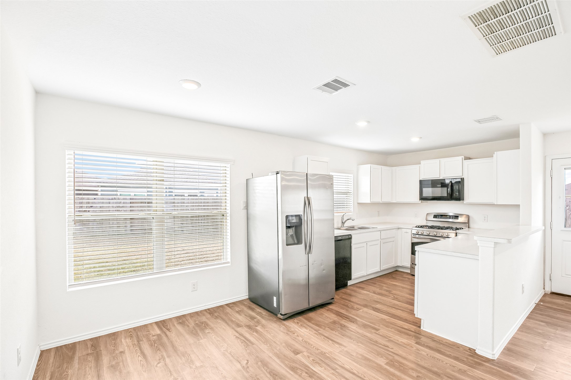 Open kitchen and dining area with white cabinetry, stainless appliances, peninsula, window blinds, and wood-look flooring.