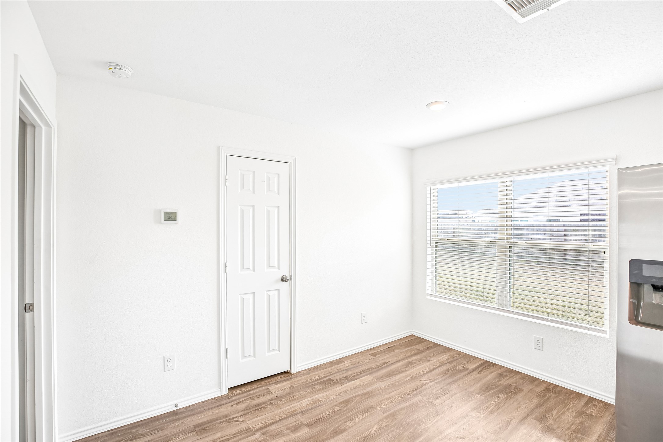 Empty kitchen/dining area with wood-look flooring, a large window with blinds, a white door, and a visible refrigerator edge.