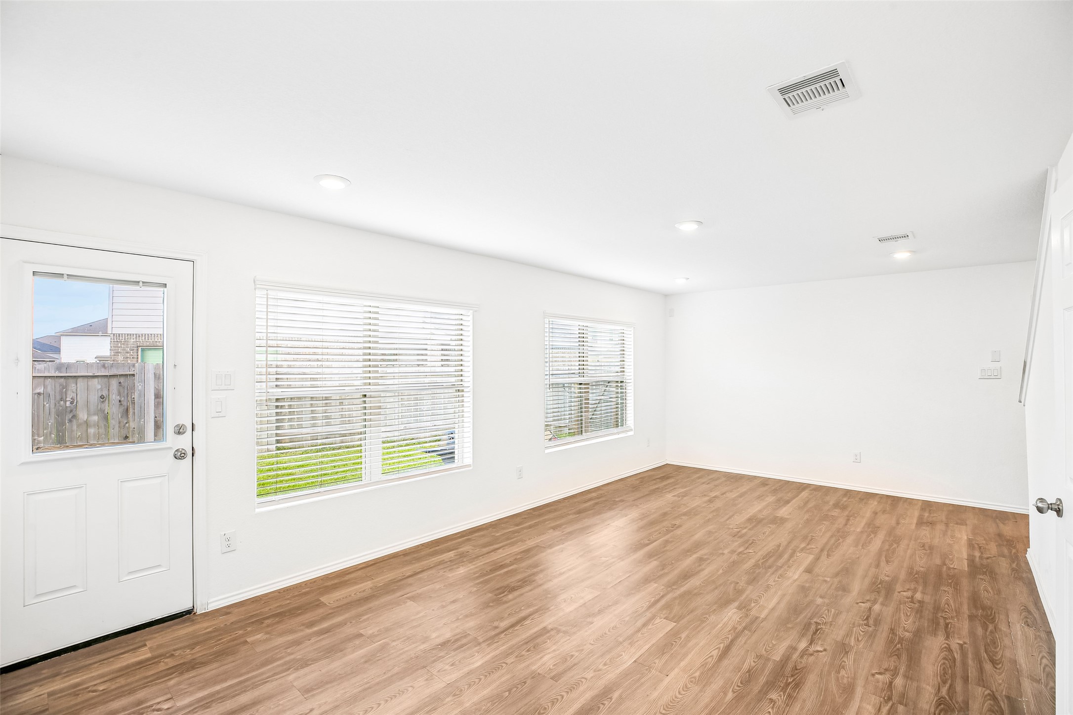 Empty living and dining area with multiple window blinds, an entry door, recessed lighting, and wood-look hard flooring.