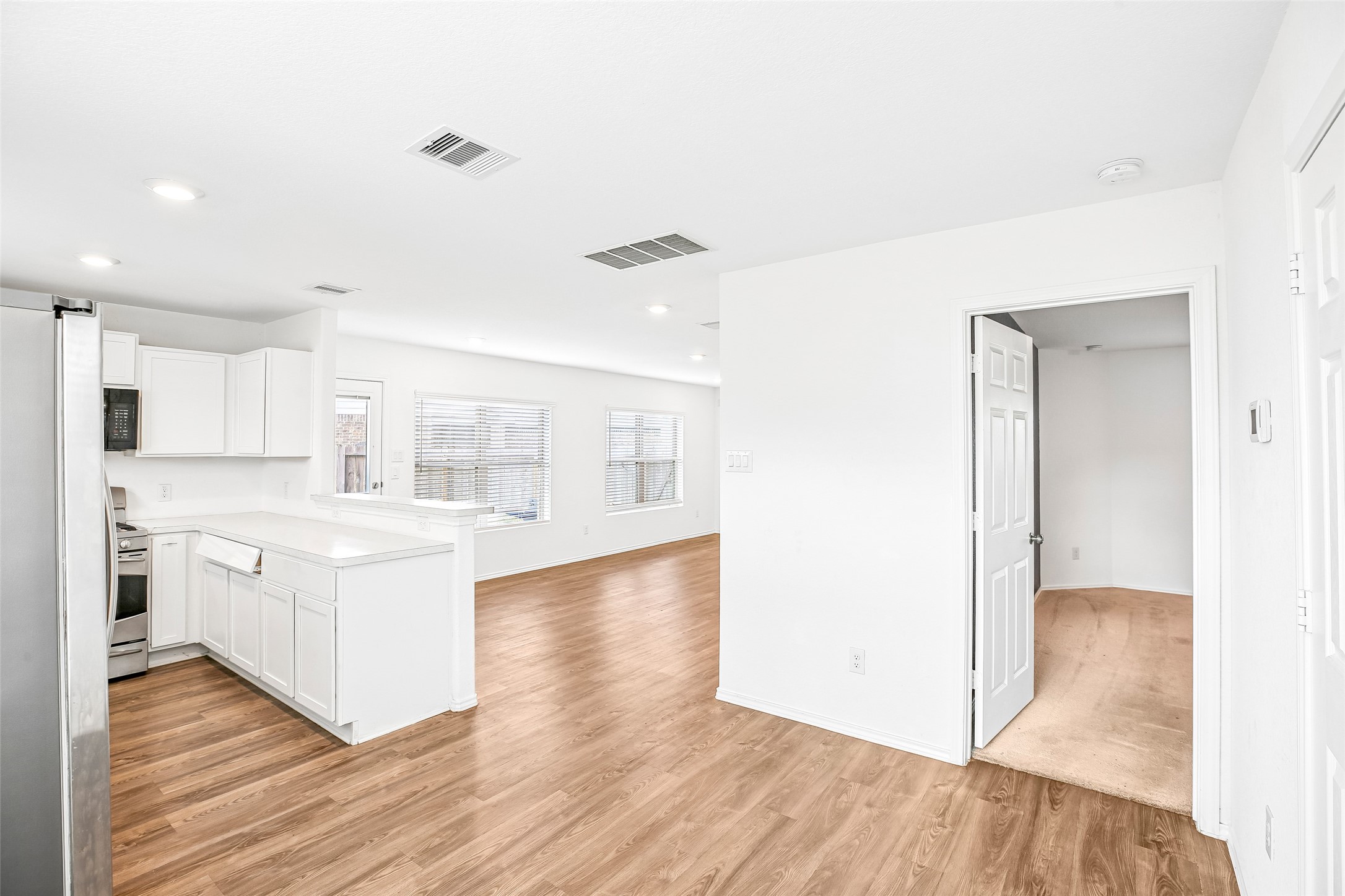 Open kitchen and dining/living area with white cabinetry, wood-look flooring, and a carpeted room beyond the doorway.