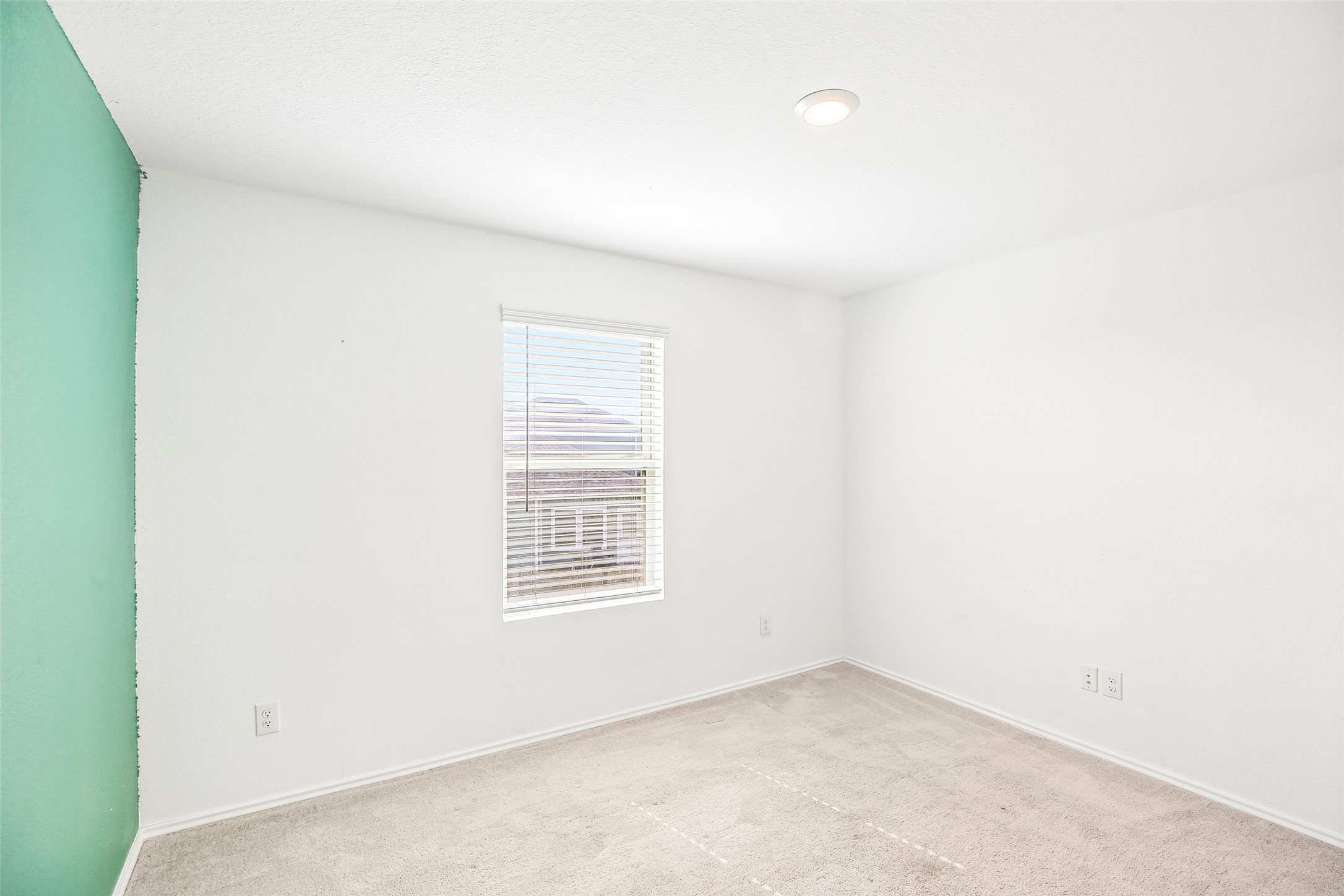 Empty carpeted interior room with a window and blinds, light walls, and a turquoise accent wall at left.