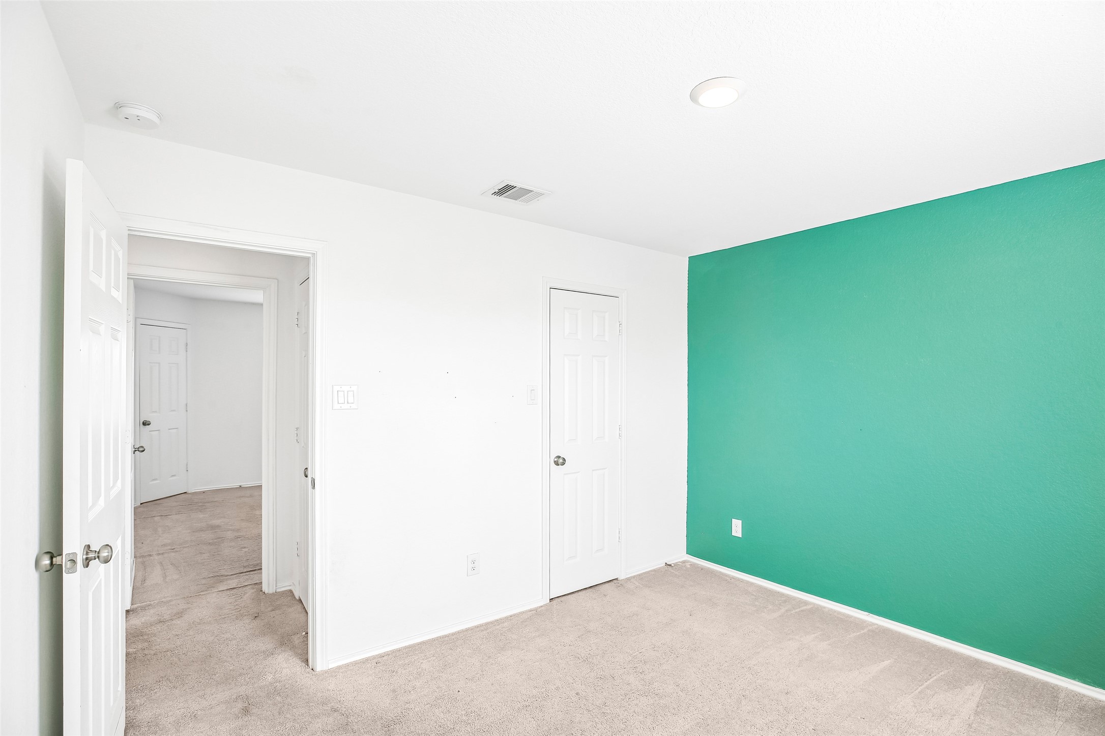 Empty carpeted interior room with a vivid teal accent wall, paneled doors, and an open hall doorway.