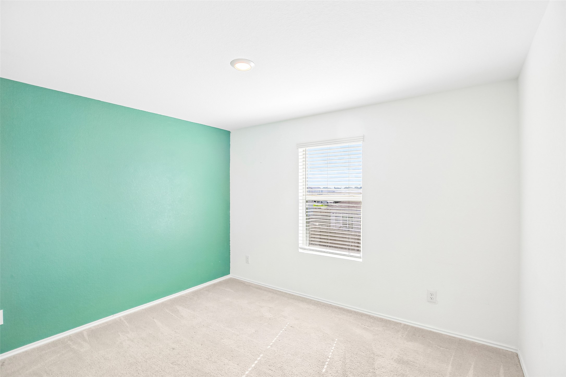 Empty carpeted bedroom with a turquoise accent wall, a window with white blinds, and a recessed ceiling light.