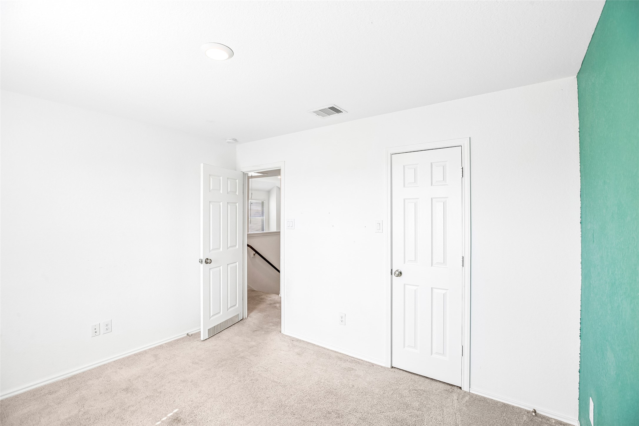 Empty carpeted bedroom with an open stairway doorway and a contrasting turquoise wall edge.