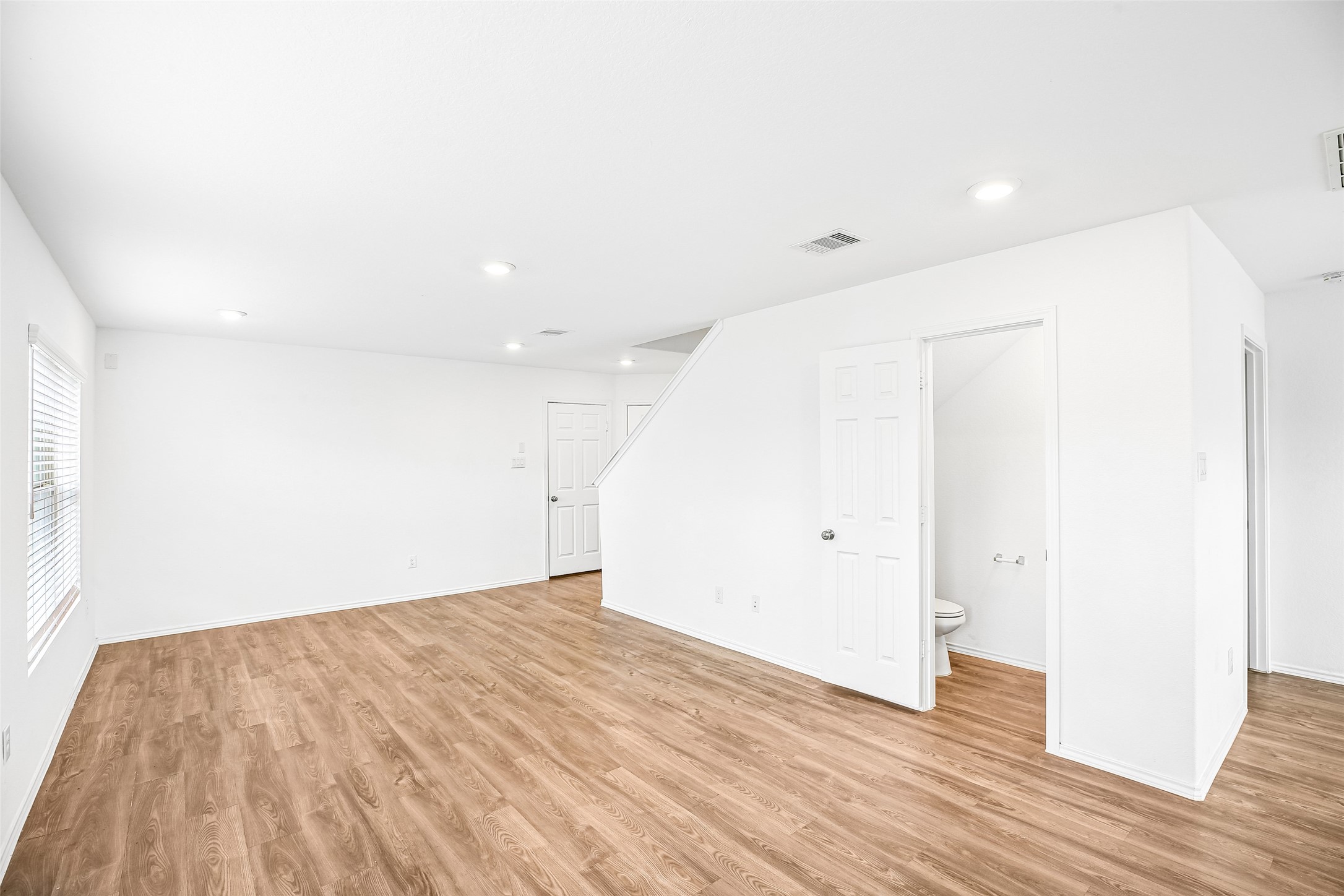 Bright, open living and dining area with wood-look flooring, a window with blinds, and a stair opening.