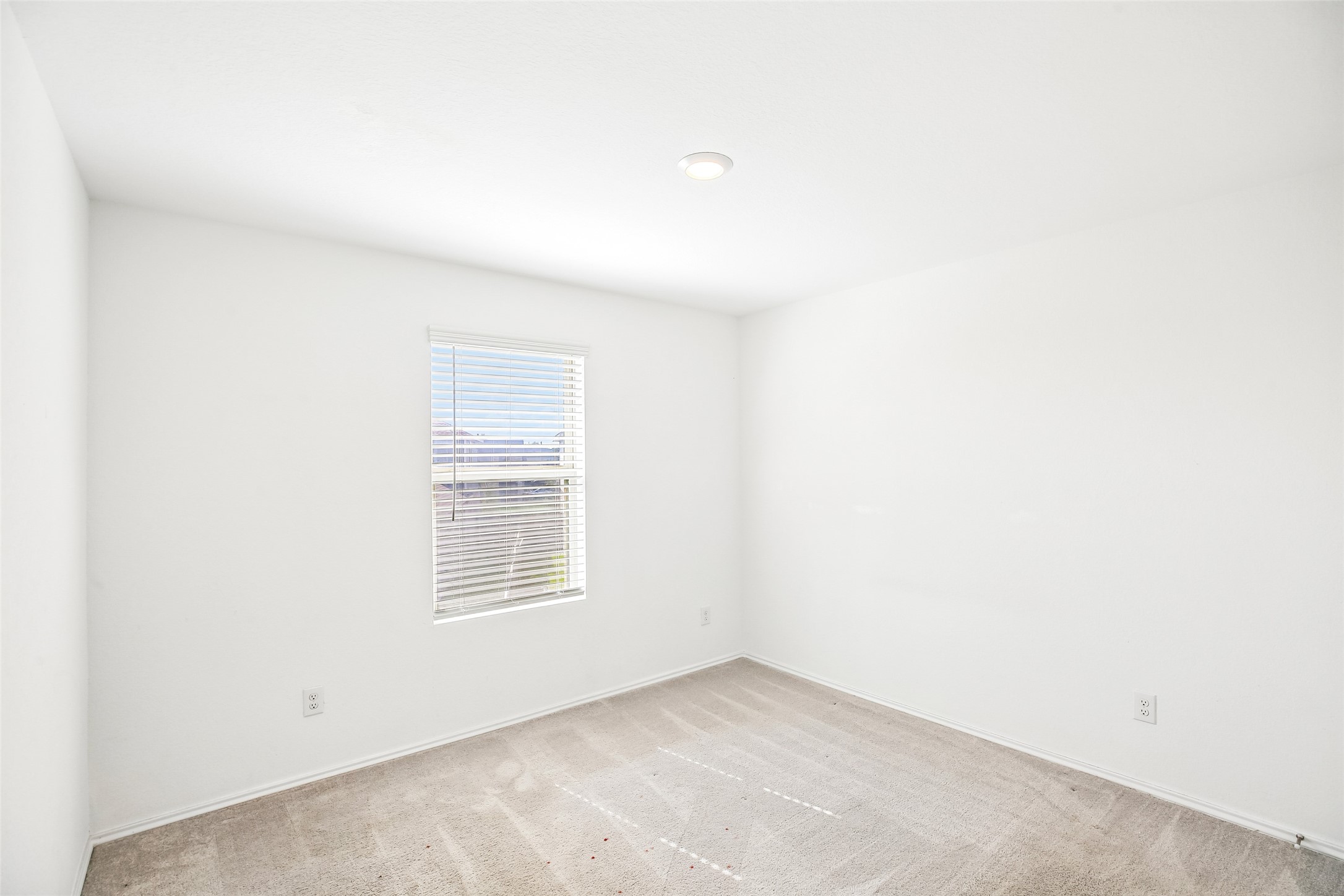 Empty carpeted interior room with a window, white blinds, recessed light, baseboards, and visible outlets.