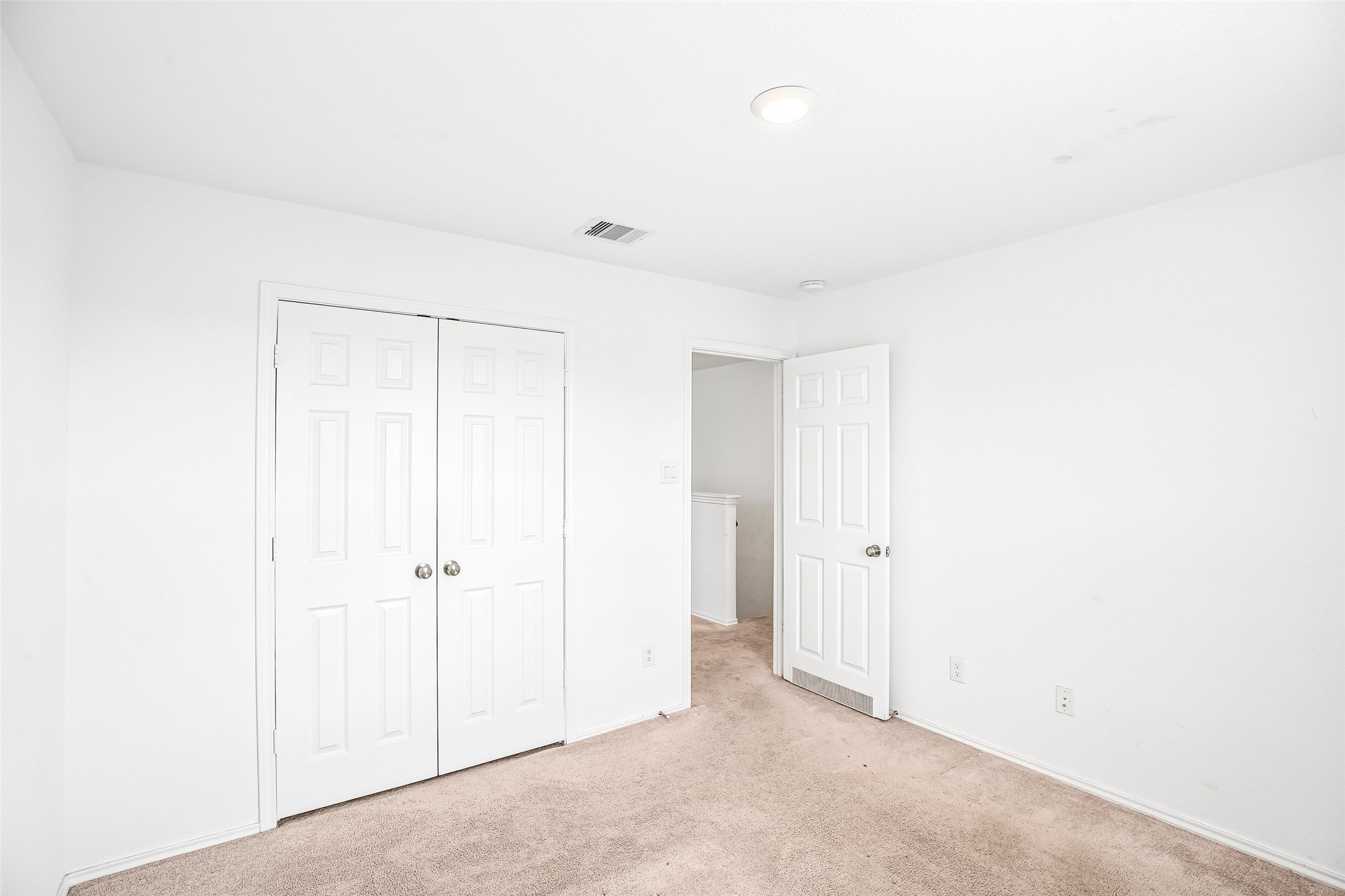 Empty carpeted interior room with double closet doors, an open hall doorway, light walls, and a ceiling vent.