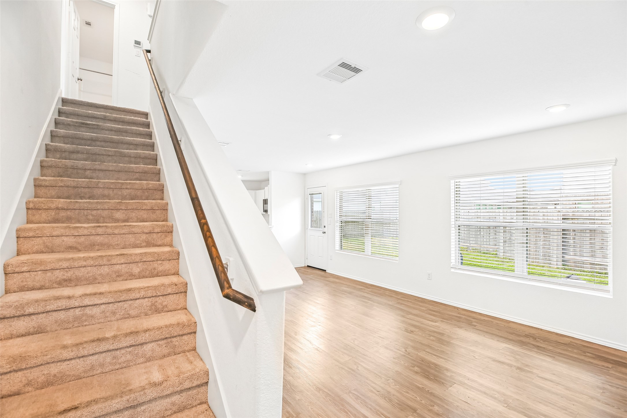 Bright, empty living/dining area with a carpeted staircase, wood-look flooring, window blinds, and entry door.