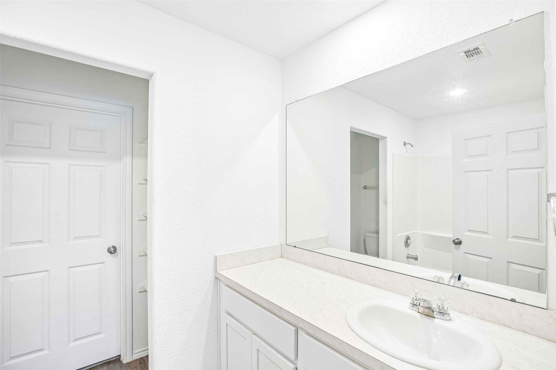 Bright bathroom view with a white vanity, large mirror, tub/shower area, white paneled doors, and dark wood-look flooring.