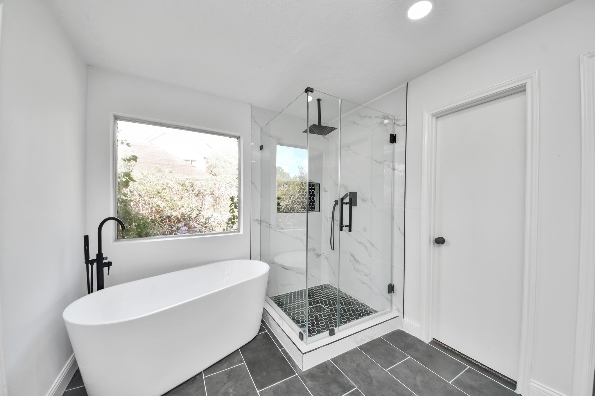Luxurious bathroom featuring a sleek freestanding tub, a modern glass-enclosed shower with elegant tilework, and abundant natural light through a large window.