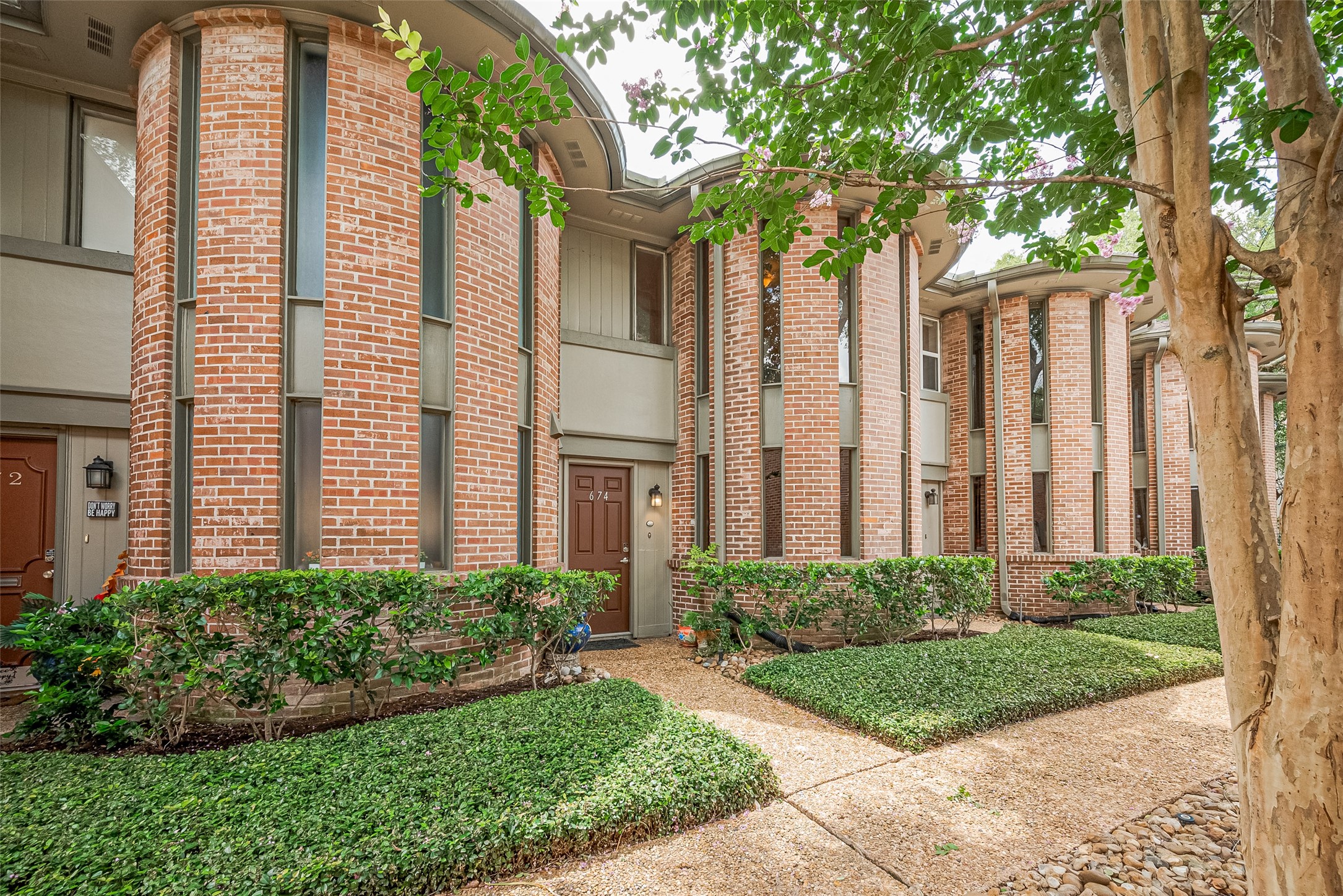 Charming townhouse features a unique curved brick facade with vertical windows, surrounded by lush greenery and a well-maintained pathway, offering a blend of modern design and natural beauty.