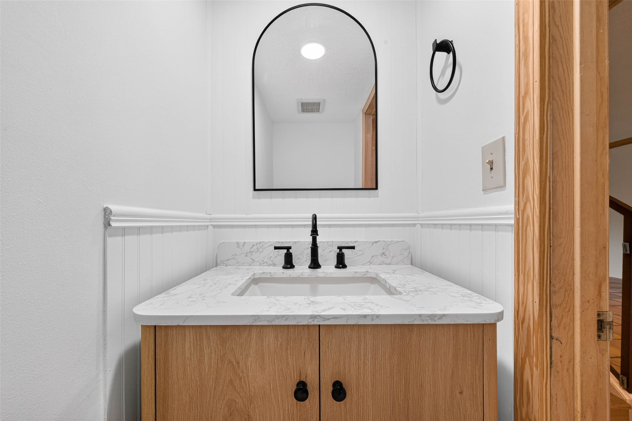 Contemporary bathroom with a sleek countertop, stylish black fixtures, and a modern arched mirror, complemented by a warm wooden vanity.