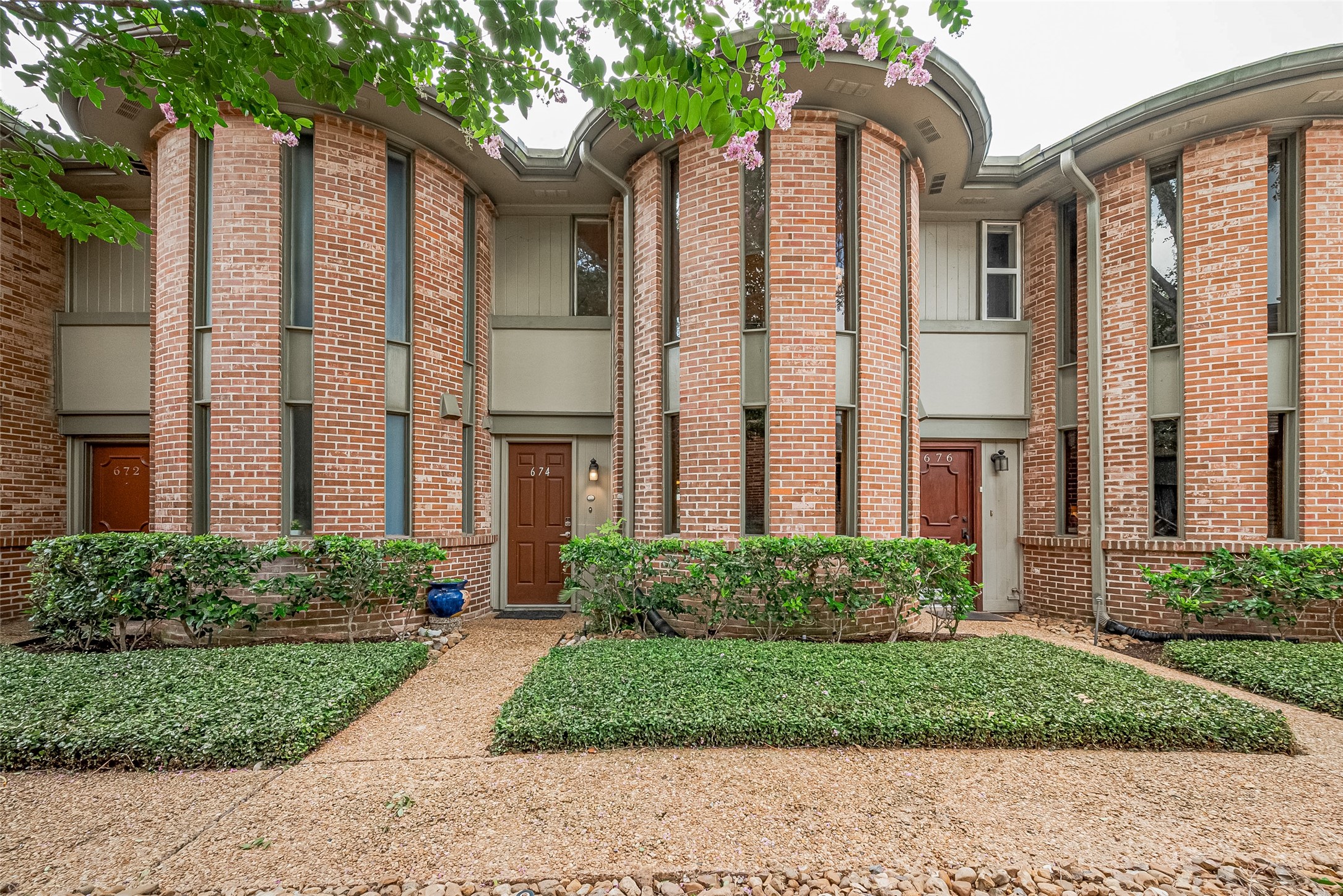 Charming brick townhouse with distinctive curved architecture and lush landscaping, offering a welcoming entrance.
