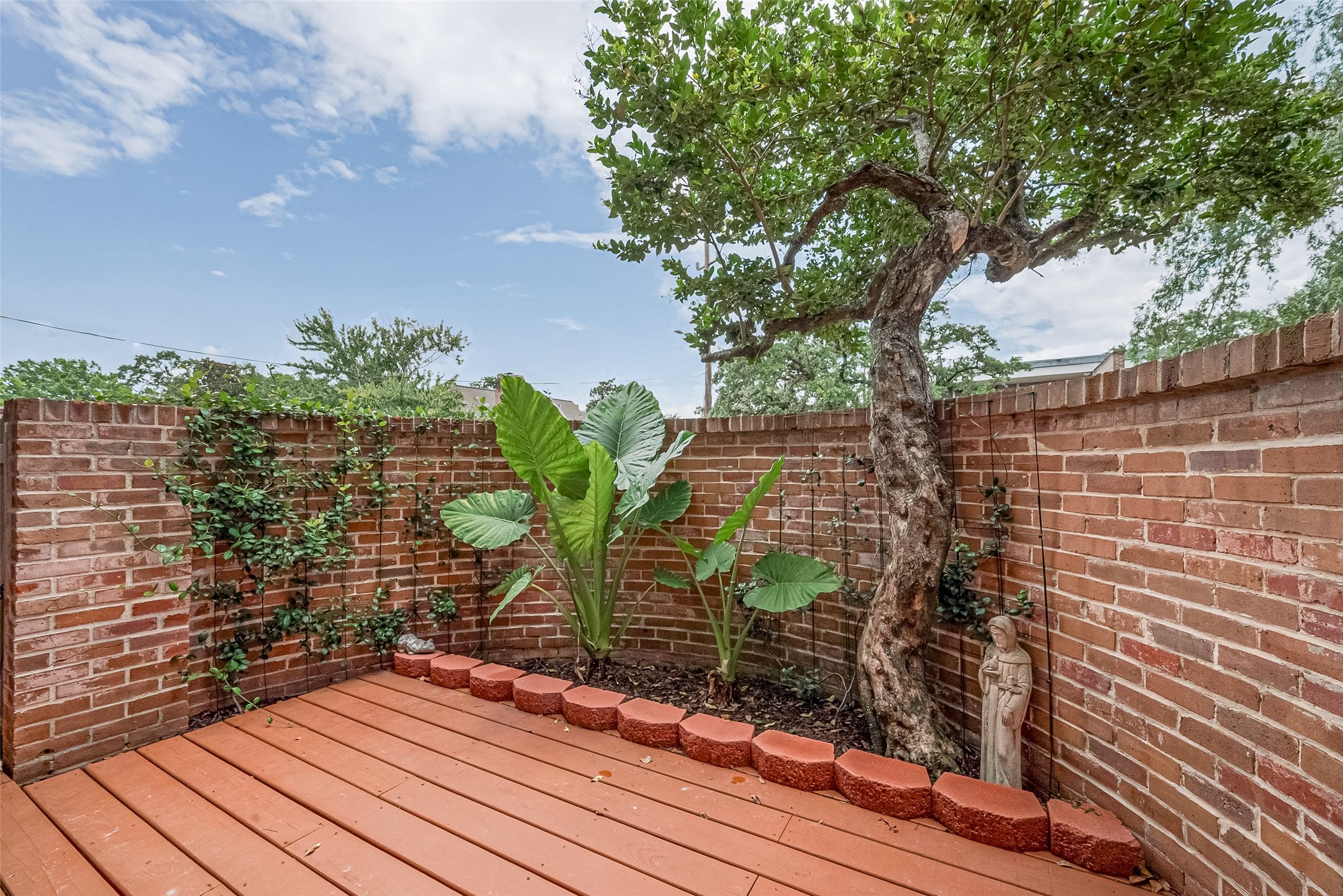 Charming private patio with a brick surround, featuring lush greenery and a mature tree for a serene outdoor retreat.