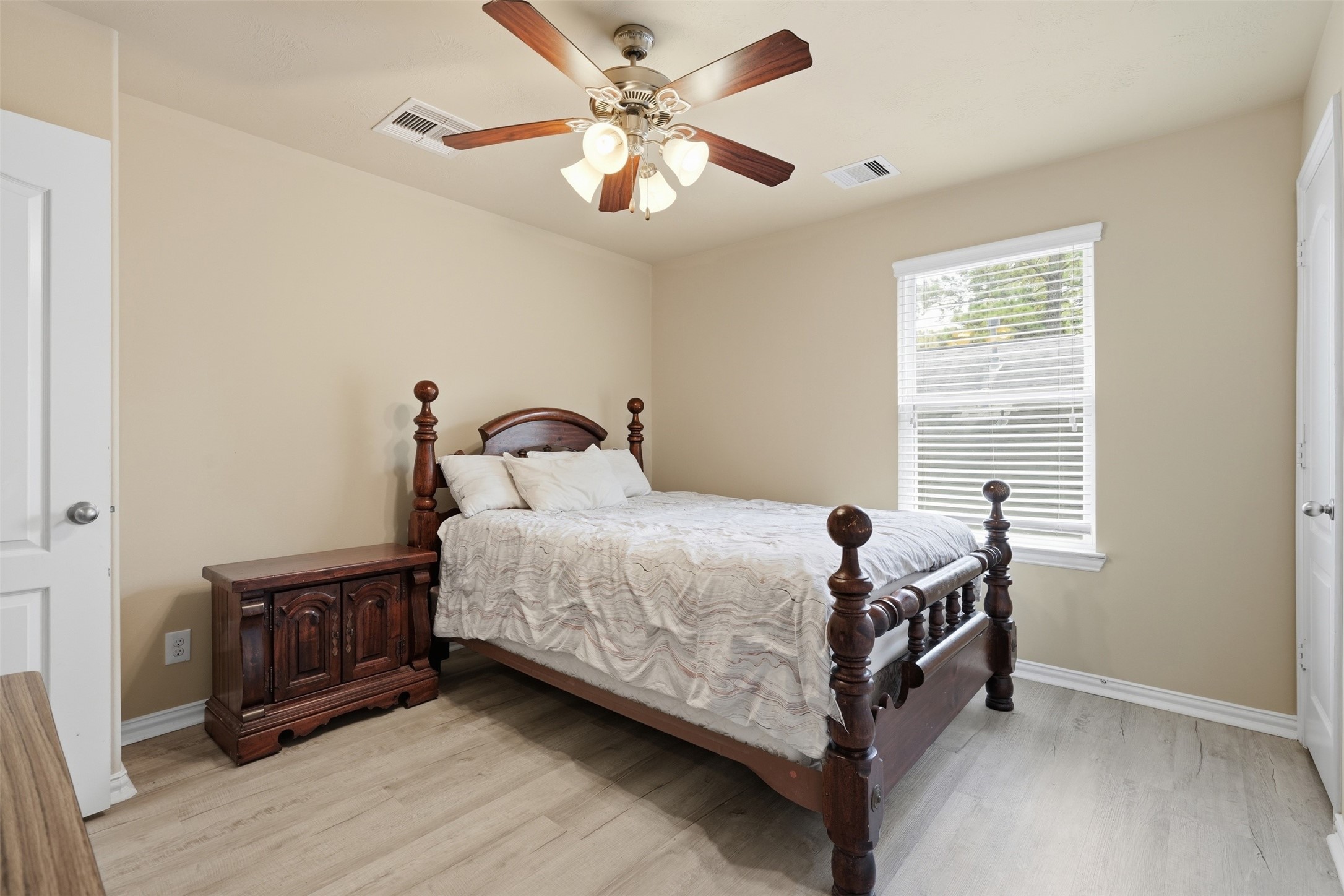 A secondary bedroom with a ceiling fan, wood-look plank floors and a window with leafy views, with room for full-size furniture.