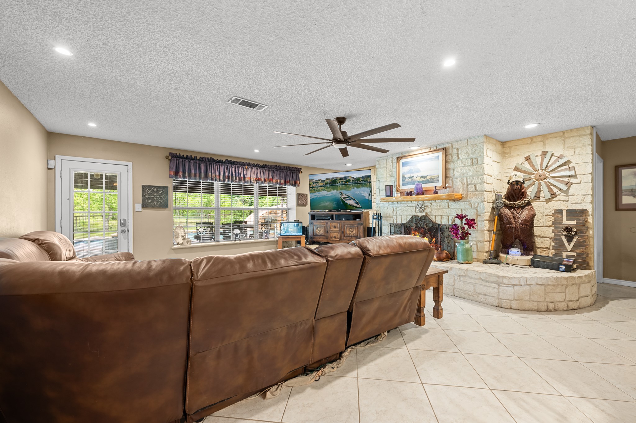 Expansive living area featuring a striking stone fireplace, recessed lighting, and oversized windows that fill the space with natural light. The open layout and generous seating area make this room ideal for relaxing or entertaining guests.