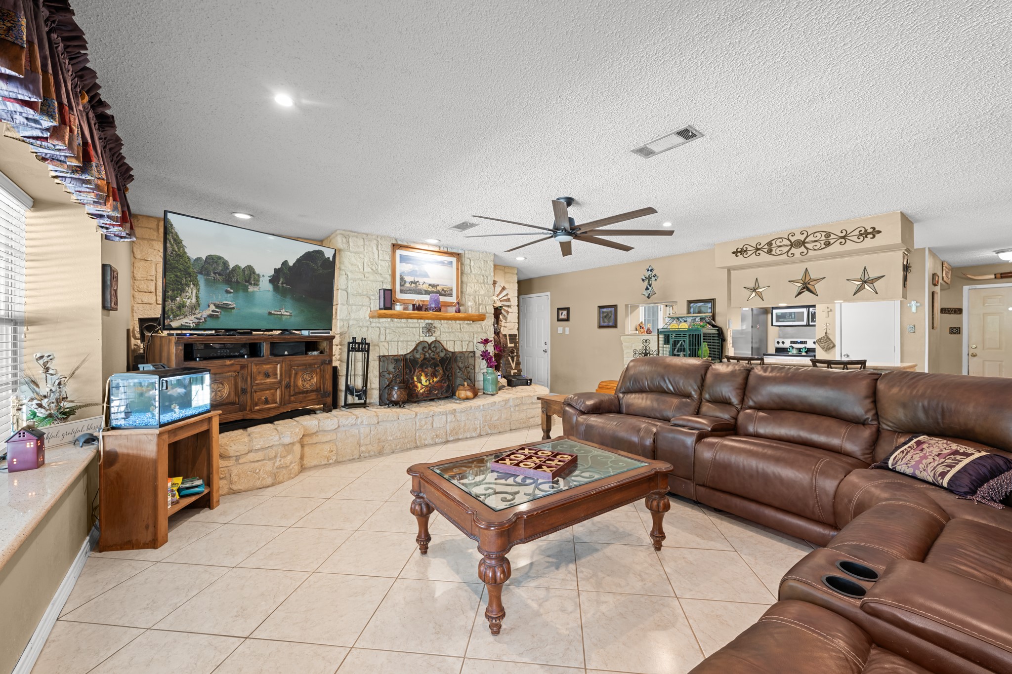 Inviting open-concept living area centered around a stunning floor-to-ceiling stone fireplace and expansive seating space. Recessed lighting, oversized windows, and seamless flow into the kitchen create an ideal environment for entertaining and everyday comfort.