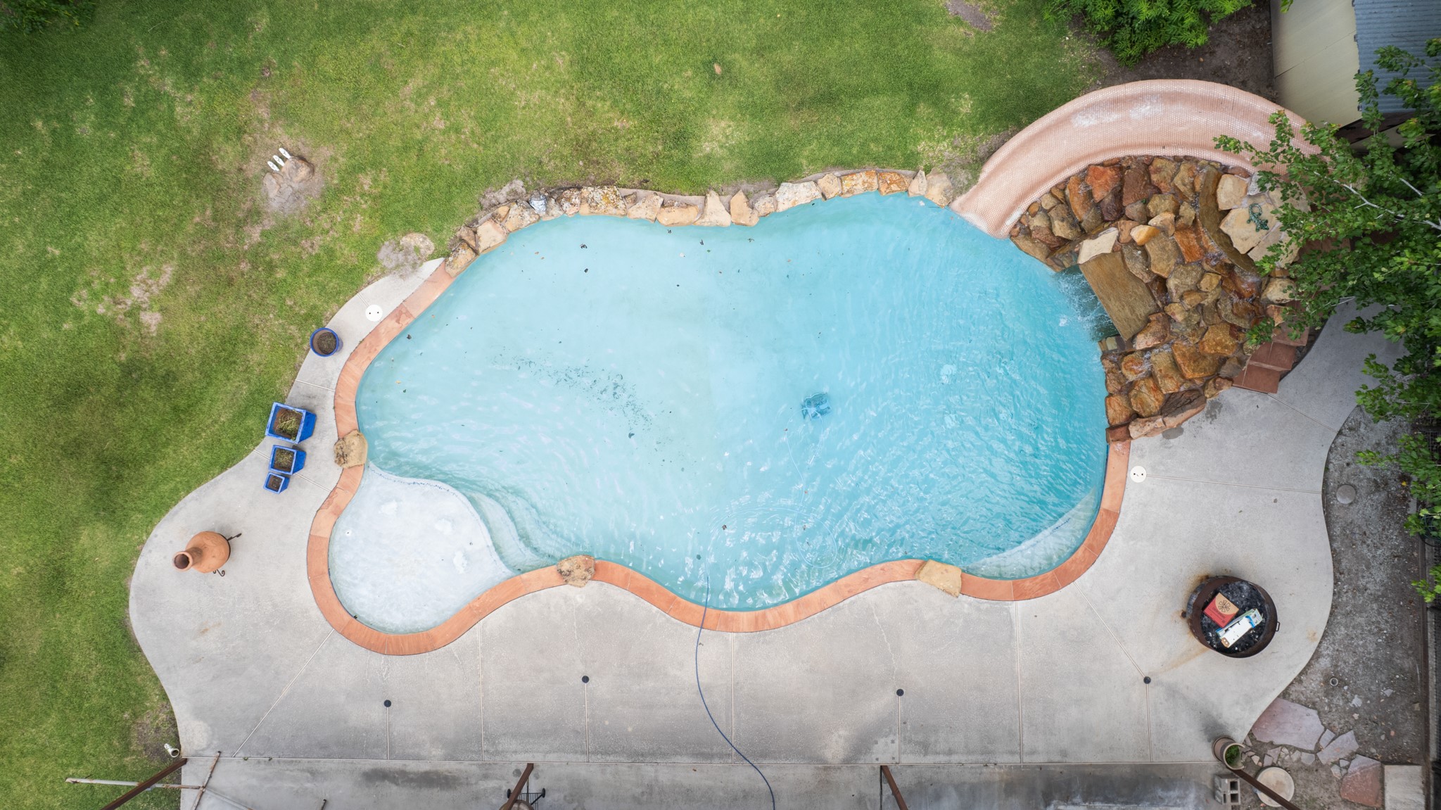 Stunning aerial view of the resort-style pool showcasing the custom rock waterfall slide, expansive concrete decking, and generous outdoor entertaining space. The freeform design and surrounding green space create a true backyard oasis.