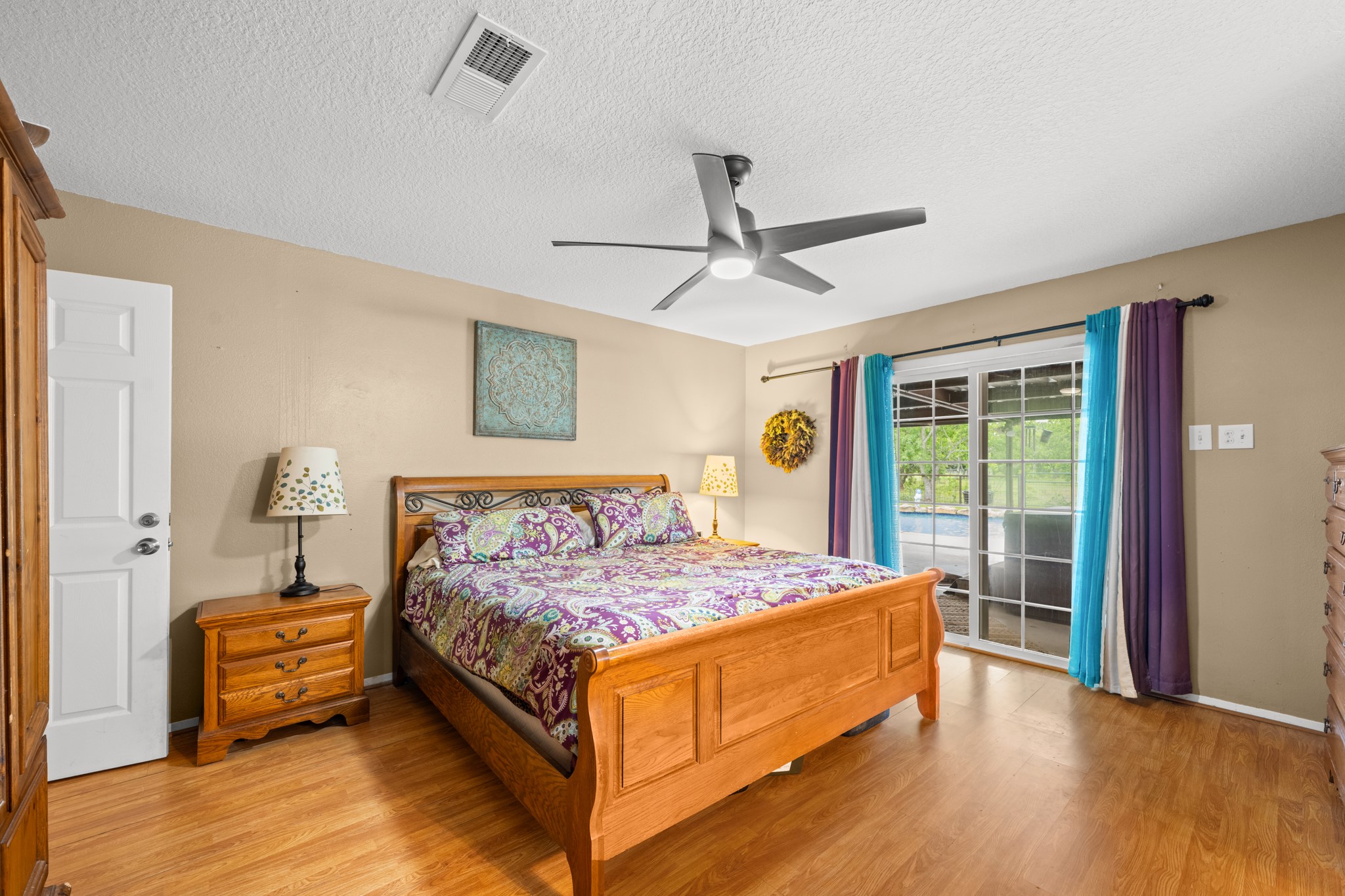Spacious primary bedroom featuring warm wood-look flooring, a modern ceiling fan, and abundant natural light. Sliding glass doors provide easy access to the outdoor living area while enhancing the rooms bright and airy feel.