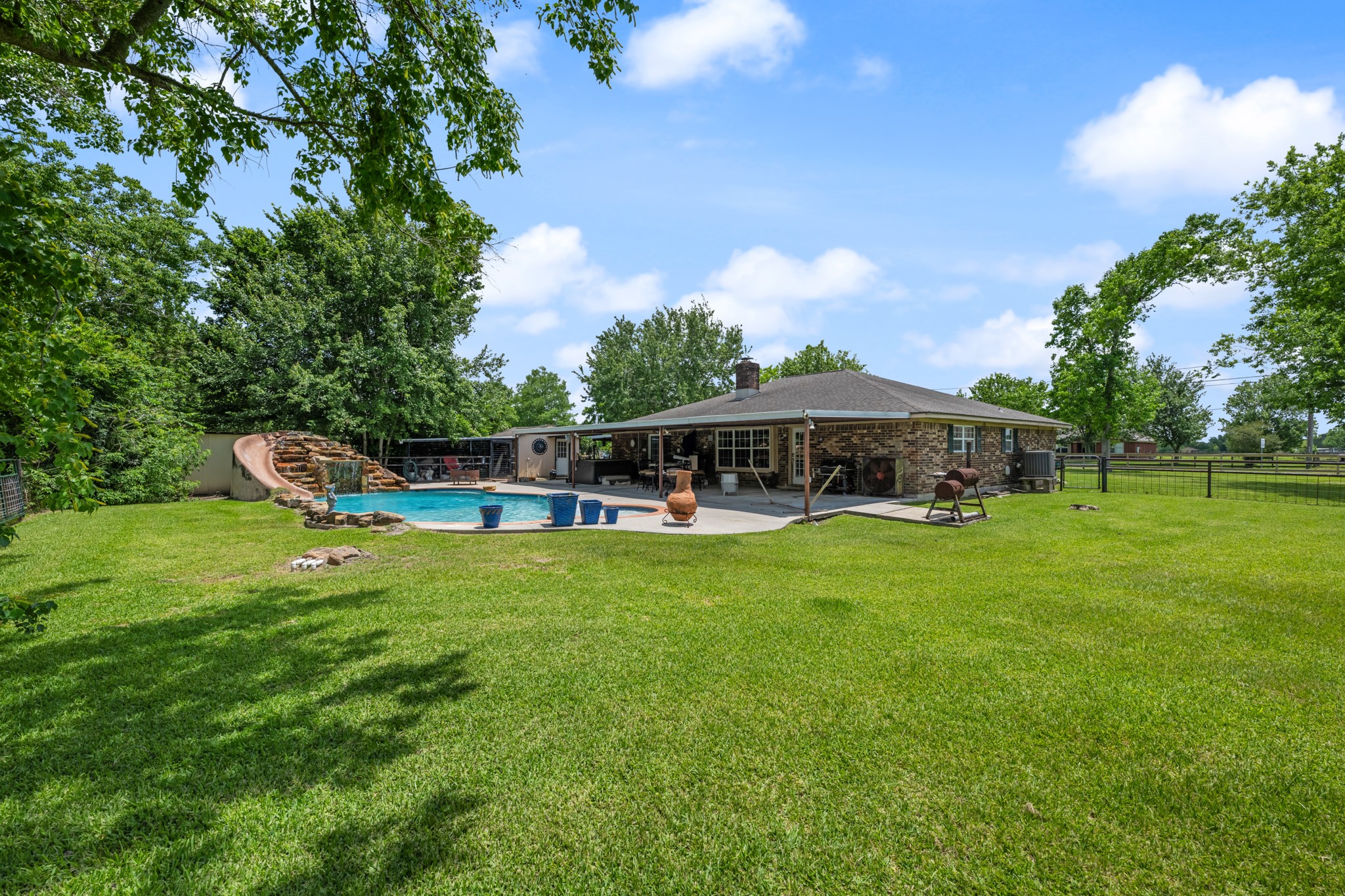 Expansive backyard retreat featuring a resort-style pool with rock waterfall slide, large covered patio, and plenty of open green space to enjoy. Mature trees and fenced surroundings create a private setting ideal for entertaining, recreation, or relaxing outdoors.
