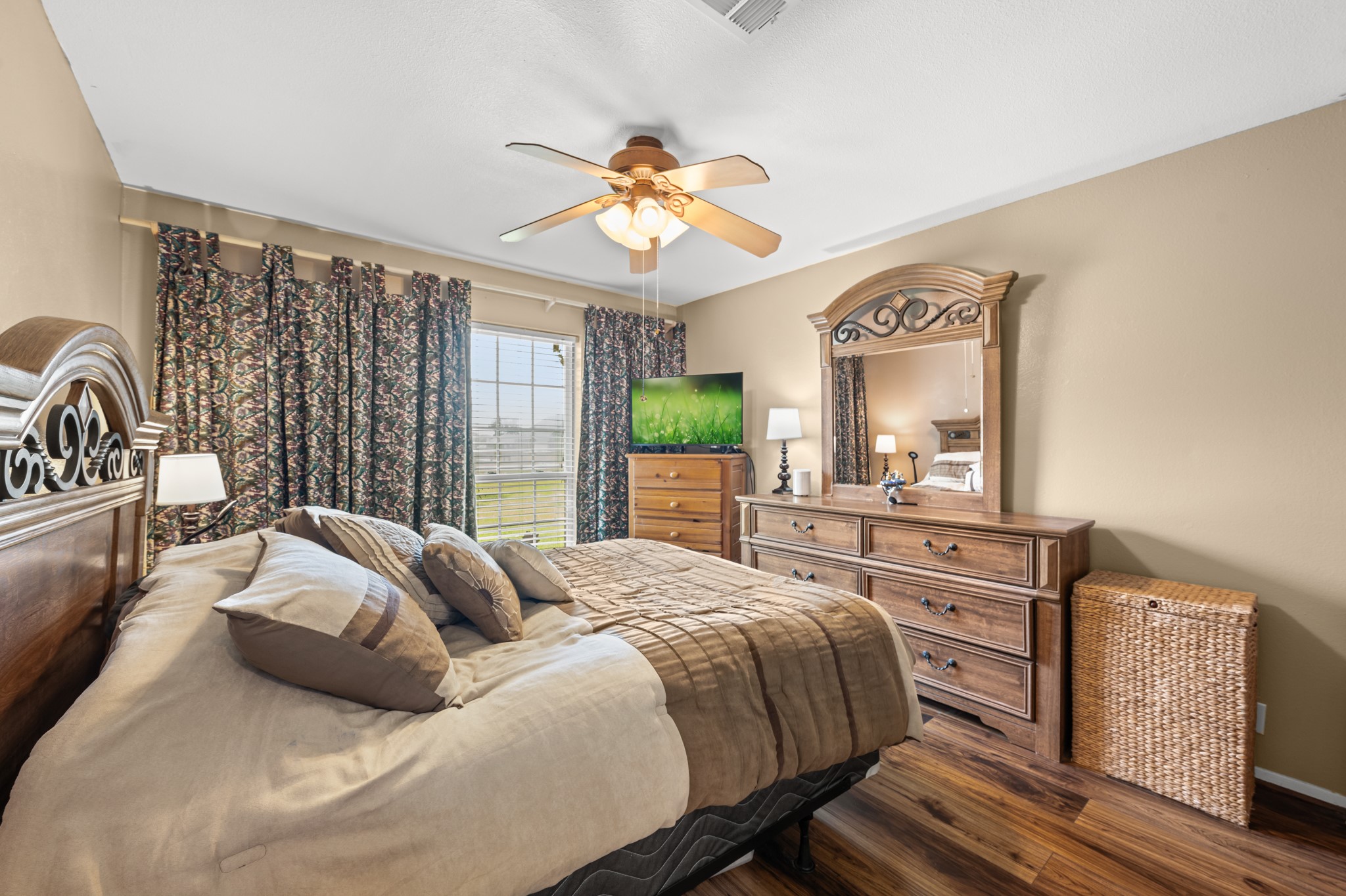 Comfortable bedroom featuring warm wood-look flooring, neutral tones, and large windows that fill the space with natural light. Generous room size and ceiling fan create a relaxing retreat with plenty of everyday comfort.
