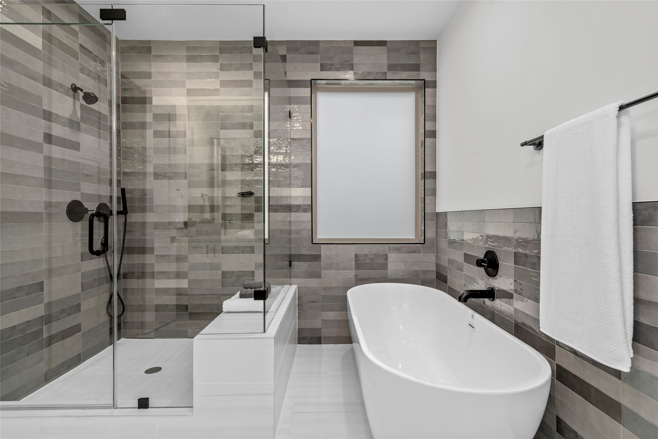 Full walls of tile surround this frameless glass enclosed shower with hand shower, contrast floor tile and a shower seat.  Floor tile is porcelain.  Another custom shade on the window over the tub.