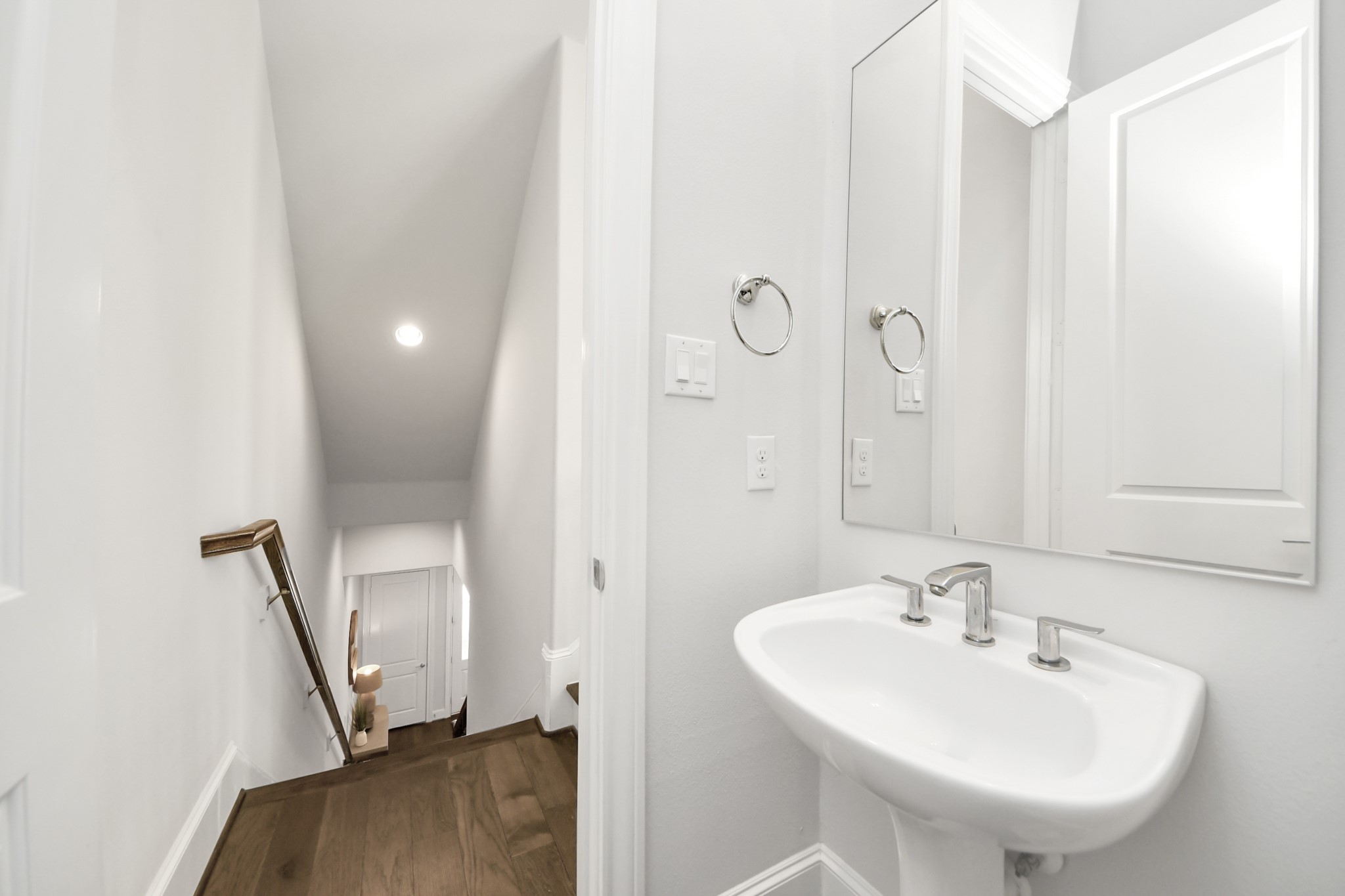 Bright and inviting hallway with elegant hardwood flooring, leading to a sleek, modern half-bath featuring a stylish pedestal sink and large mirror.
