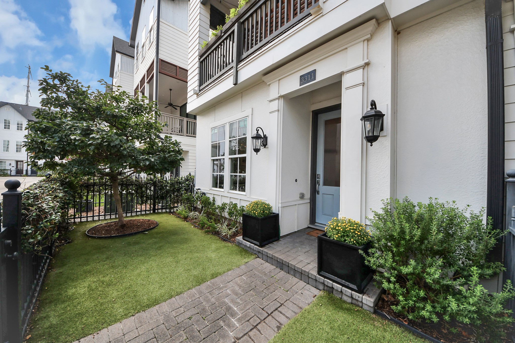 Charming entryway with a light blue door, framed by elegant lanterns and lush landscaping, leading to a quaint fenced yard with mature trees and greenery.