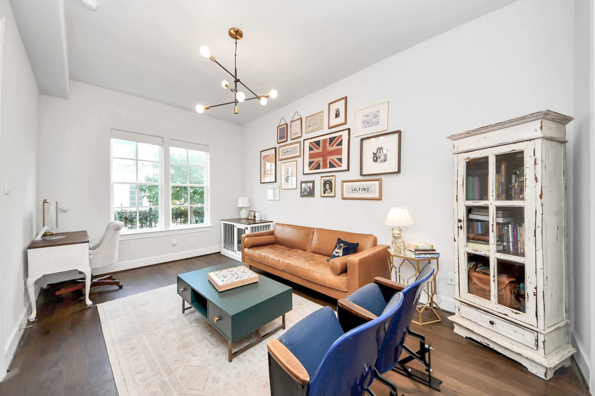 Charming living space with a warm leather sofa, eclectic wall art, and a vintage cabinet, all bathed in natural light from large windows.