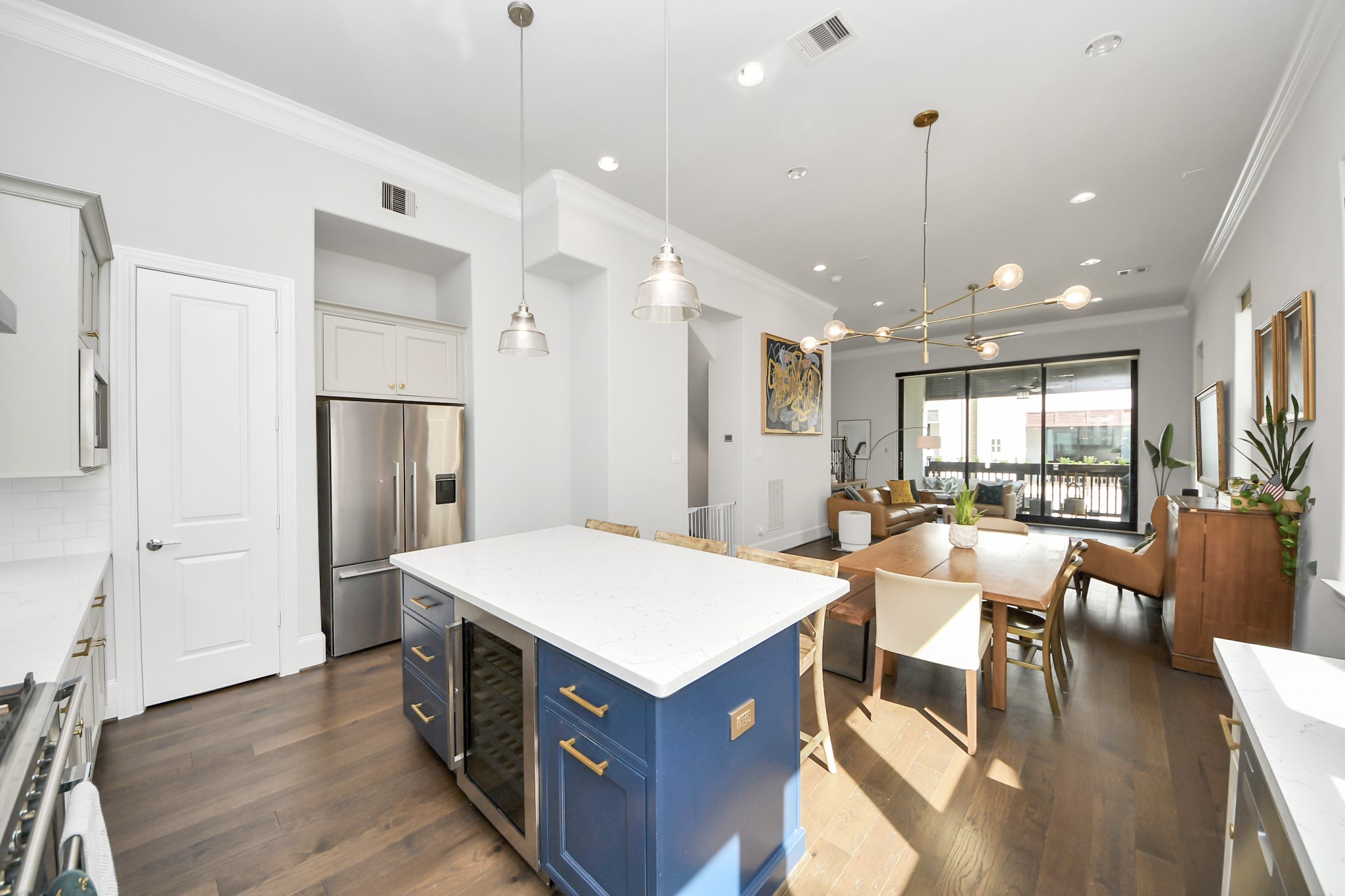 Bright and open kitchen and living area featuring a spacious island with sleek blue cabinetry, modern pendant lighting, and a seamless flow to a cozy seating area with large windows and abundant natural light.