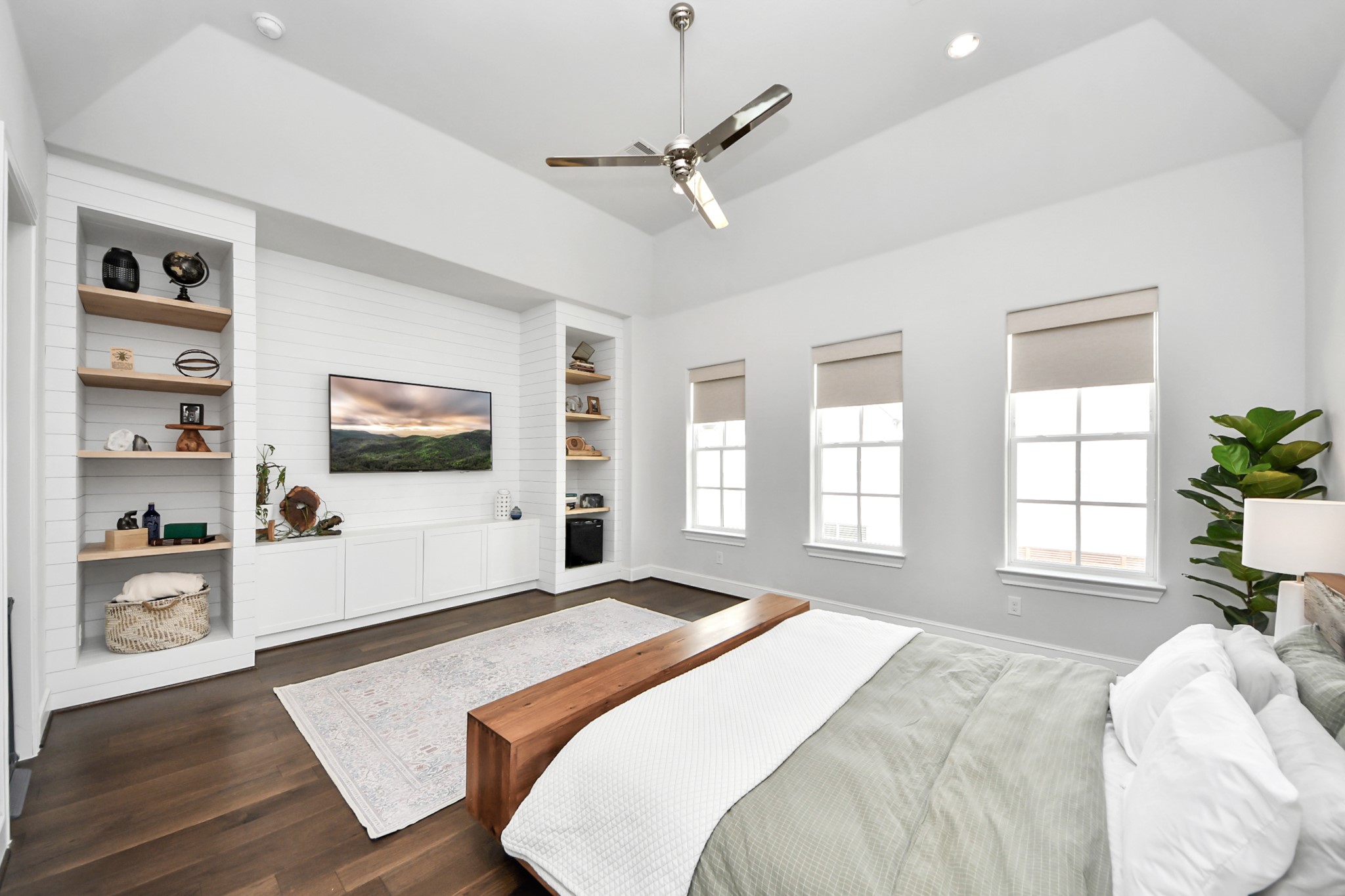 Spacious and bright bedroom featuring a vaulted ceiling, modern fan, and built-in shelving with a sleek entertainment center. Large windows allow natural light to illuminate the elegant hardwood floors and contemporary decor.