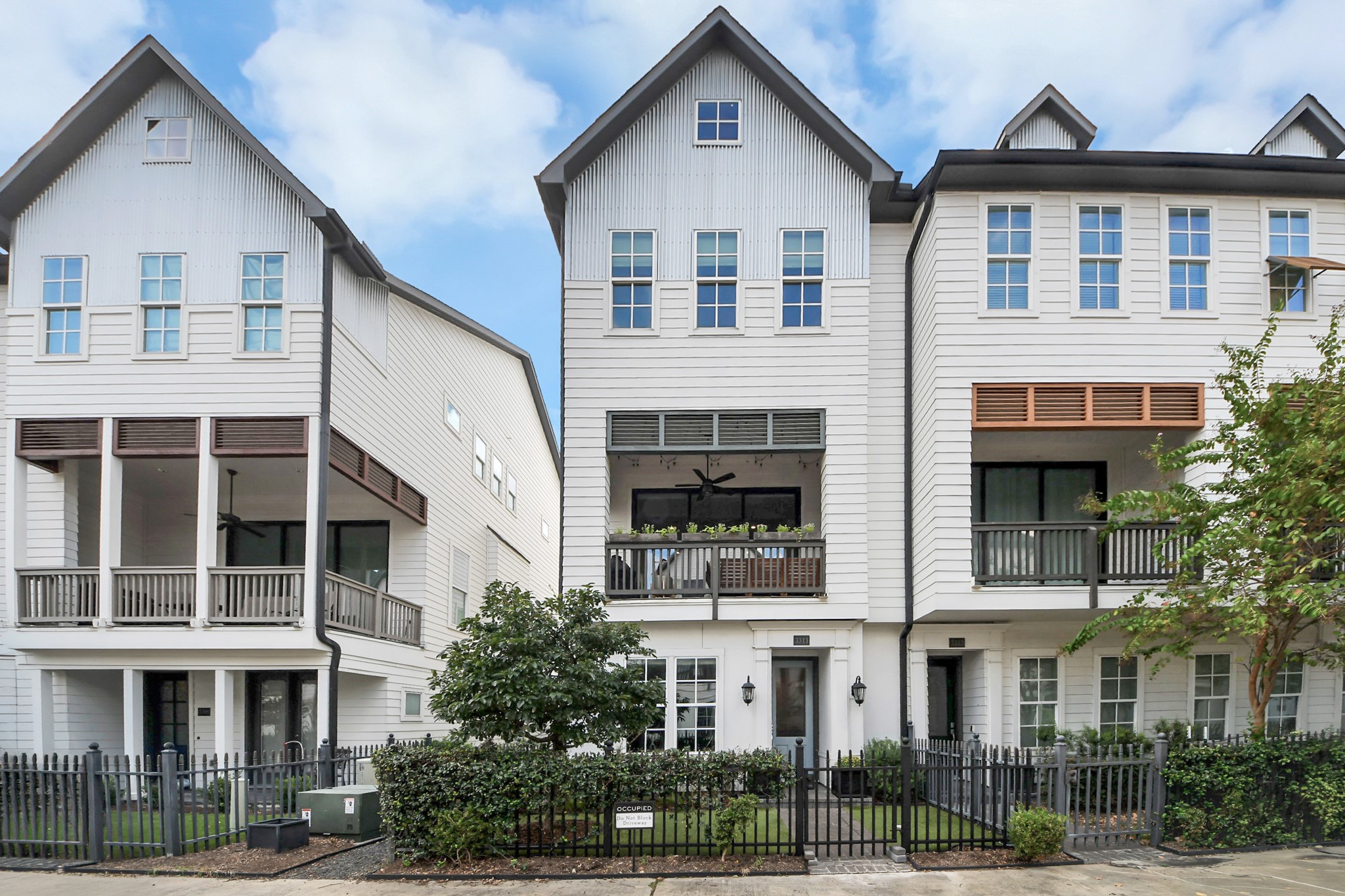 Charming three-story townhomes feature modern architecture with large windows, inviting balconies, and a neatly landscaped front yard, all enclosed by a stylish wrought-iron fence.