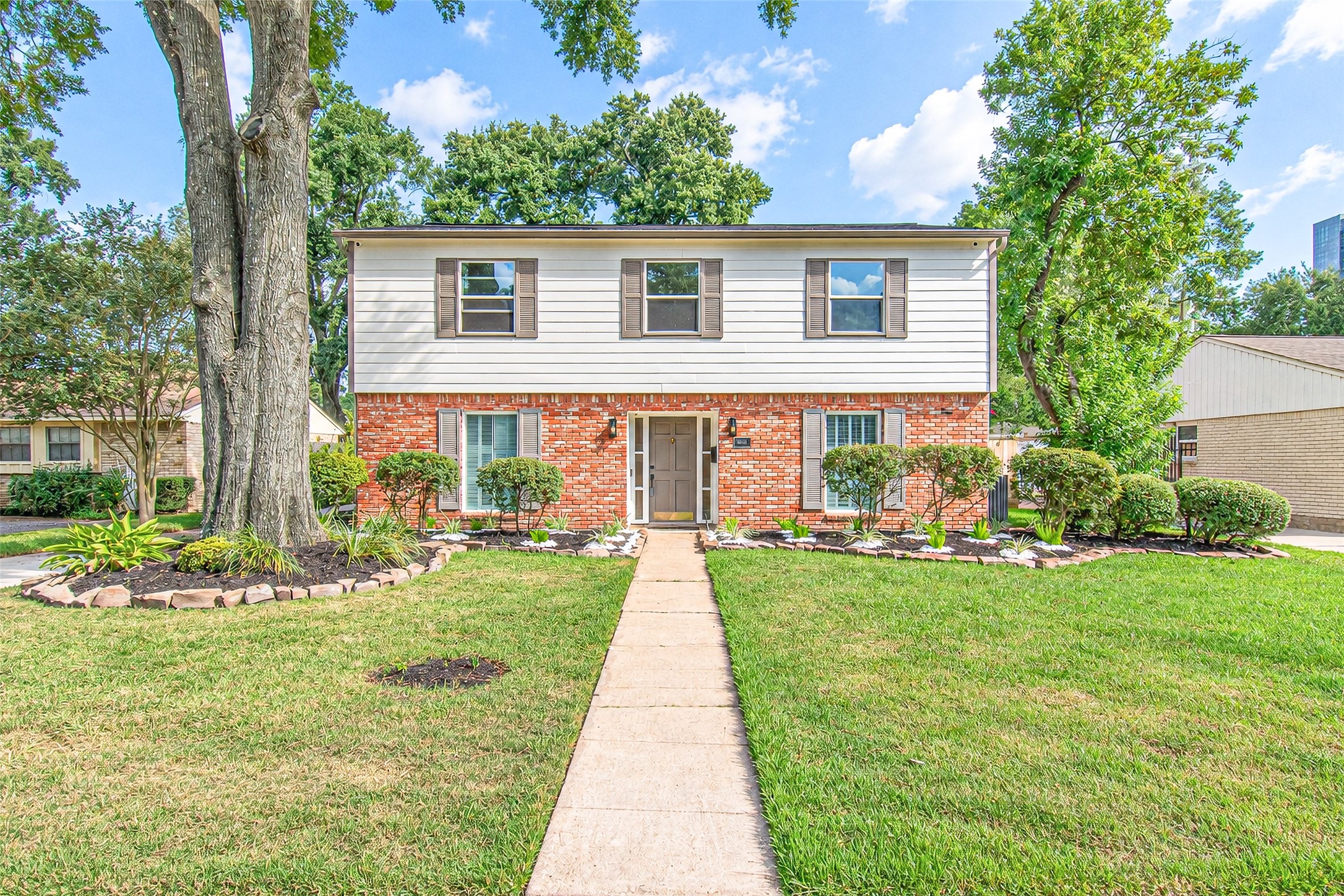 Charming two-story home with a classic brick facade and crisp white siding, surrounded by lush landscaping and mature trees, offering a welcoming pathway to the entrance.
