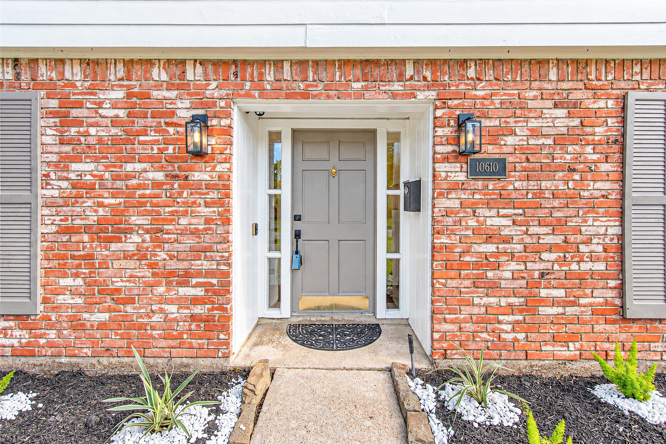 Charming brick facade with modern gray door and sidelights, framed by tasteful landscaping and elegant lighting, creates a welcoming entryway.