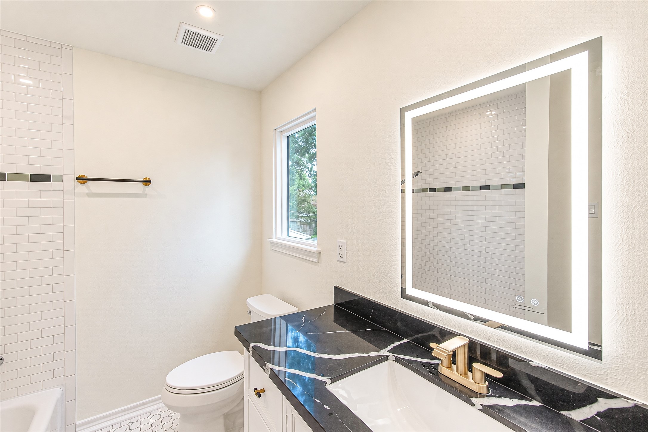 Luxuriate in the primary bath featuring a sleek black quartz countertop, modern gold fixtures, and an elegant backlit mirror, all complemented by pristine white tiles.