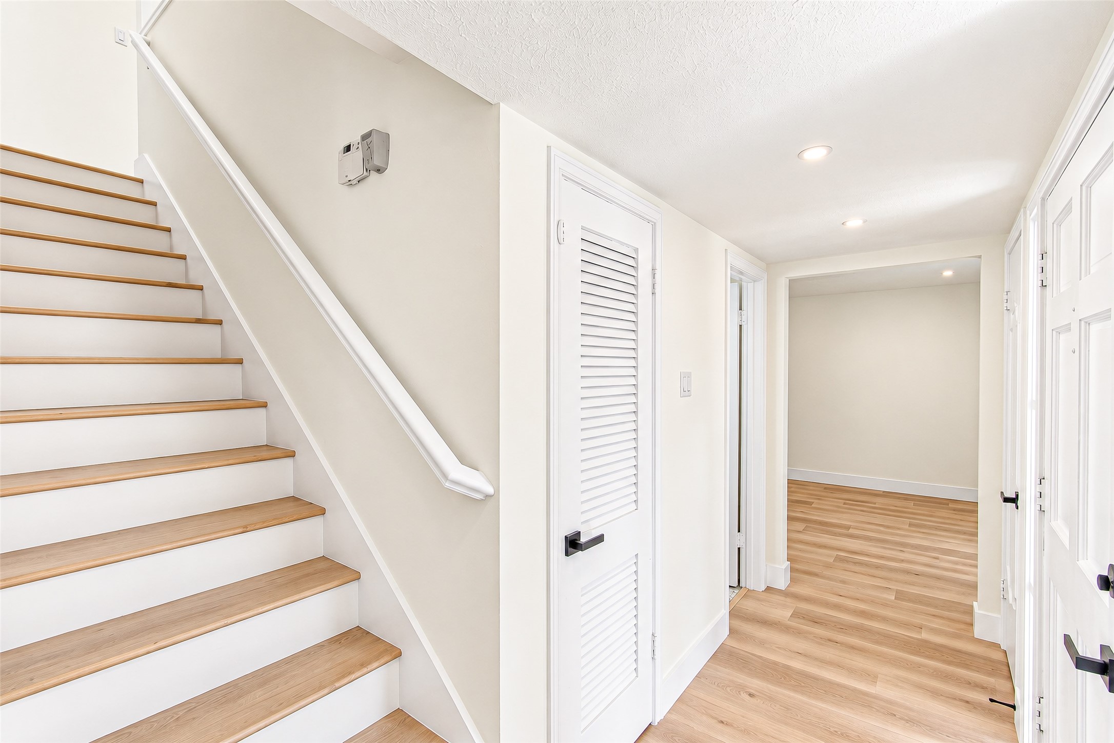 Bright and inviting hallway featuring sleek vinylflooring, crisp white walls, and modern recessed lighting leading to multiple rooms and an elegant staircase.
