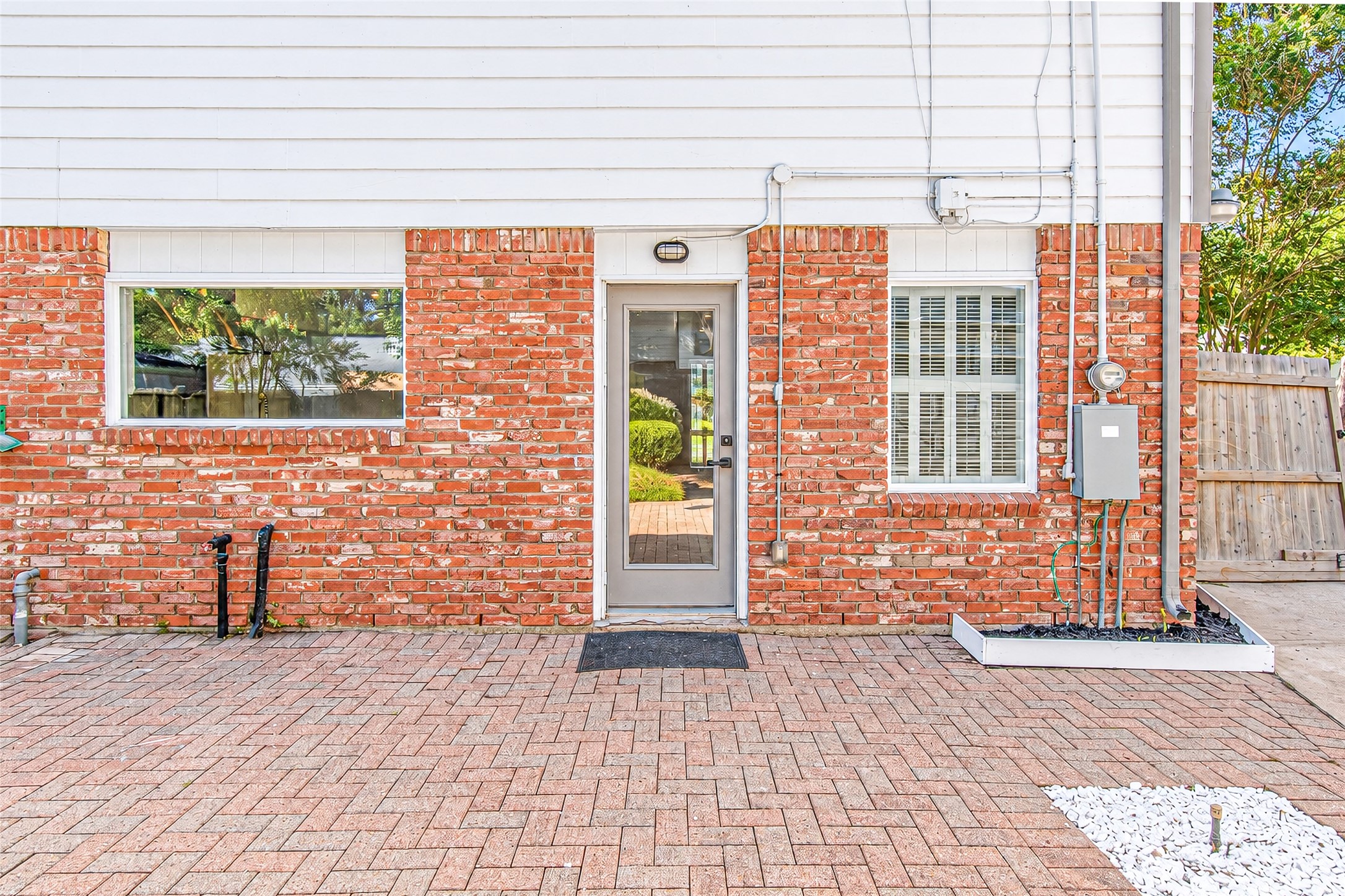 Charming brick exterior with a modern glass door, flanked by large windows for ample natural light, opens to a spacious paved patio perfect for outdoor living.