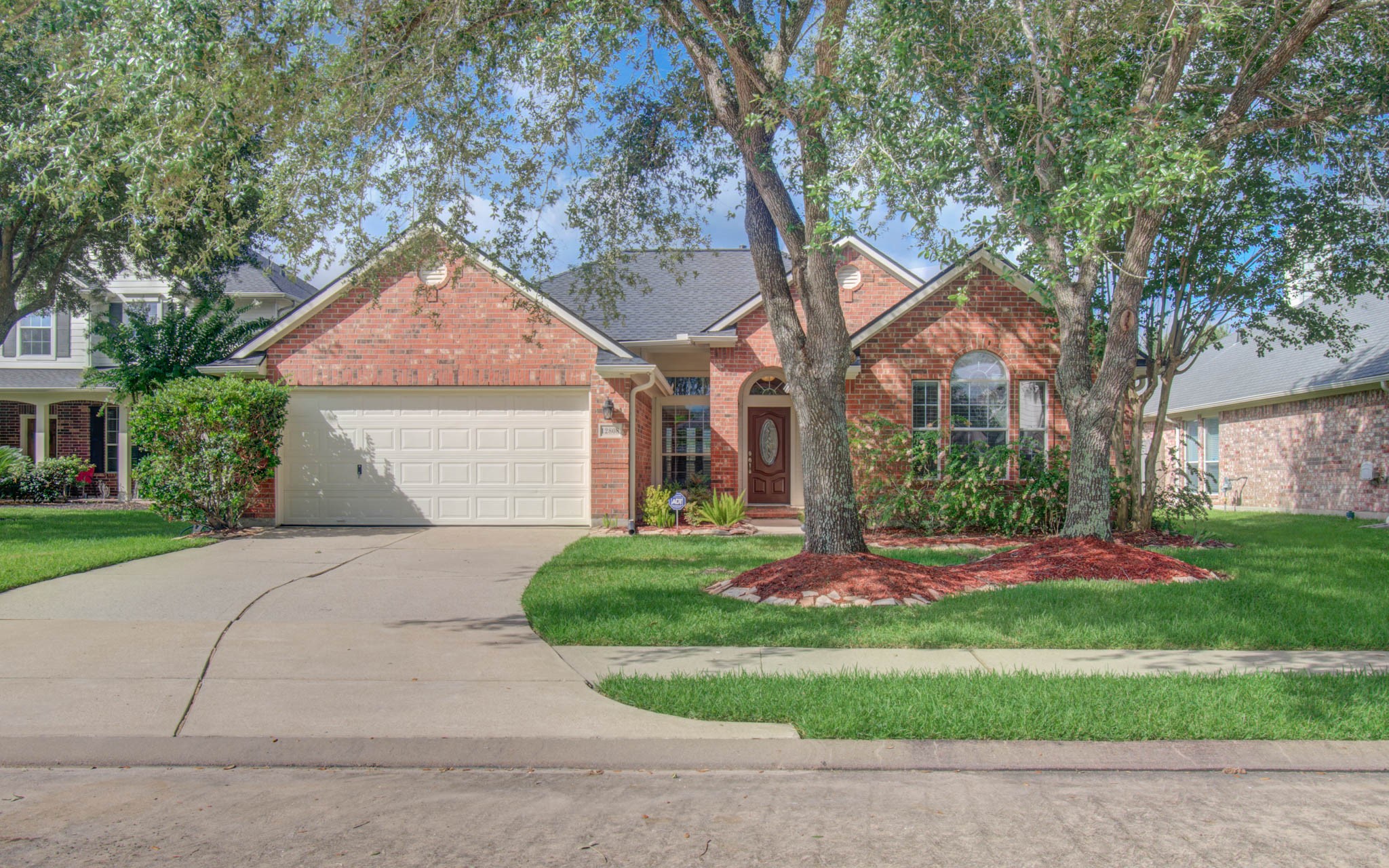 Charming brick-front home with a two-car garage nestled among mature trees, featuring a well-manicured lawn and inviting curb appeal.