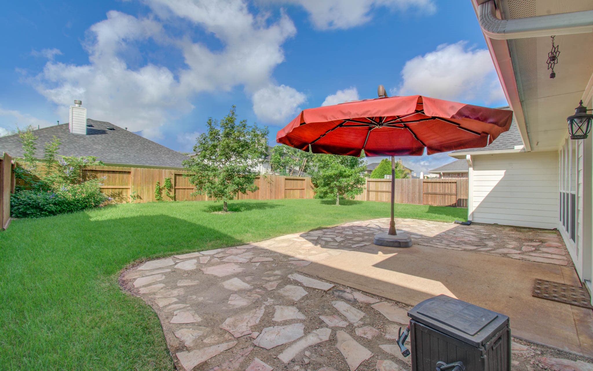 Spacious backyard features a charming stone patio with a large umbrella, perfect for outdoor gatherings and relaxation.