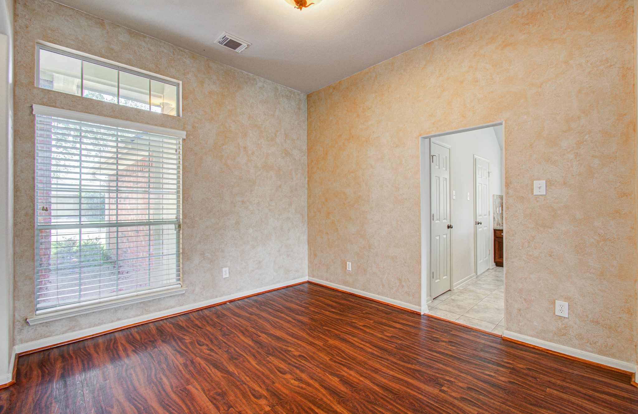 Inviting open space with rich wood flooring, a large window for natural light, and a warm, textured wall finish, leading to the kitchen