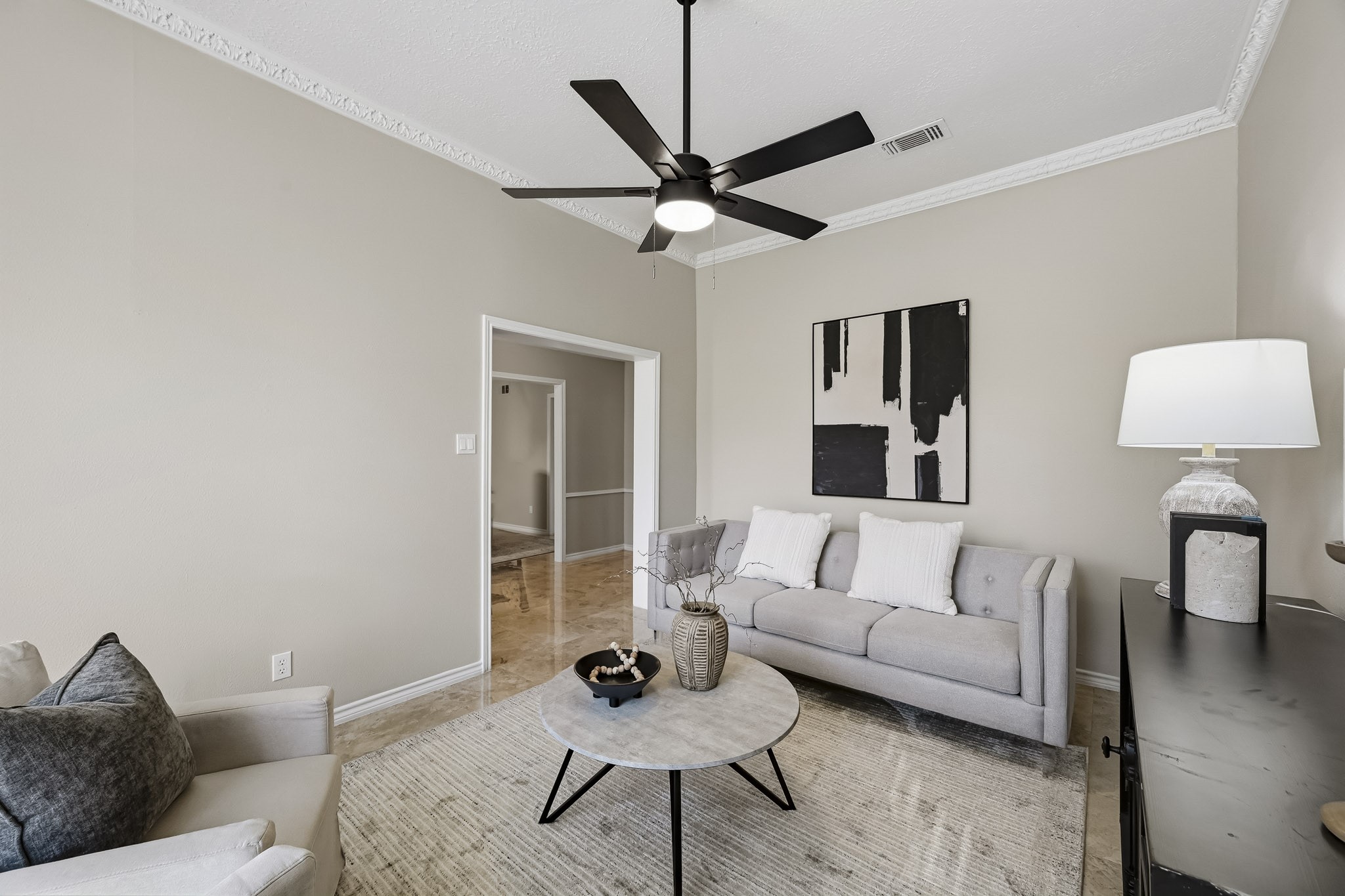 Charming living room with modern ceiling fan, neutral walls, and elegant crown molding. Cozy seating area includes a stylish sofa and armchair, complemented by contemporary decor and soft lighting.
