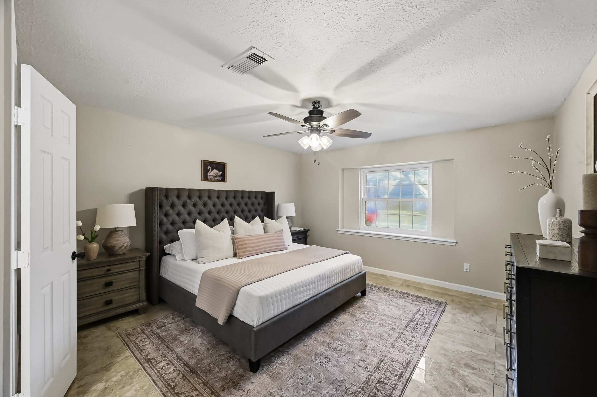 Spacious bedroom featuring a plush, tufted bed and elegant dcor, with a large window allowing natural light to enhance the warm, welcoming atmosphere.