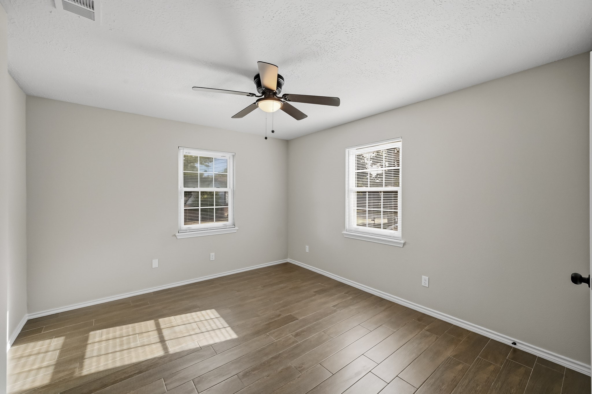 Bright and inviting bedroom featuring sleek wood-look tile flooring, neutral walls, and a modern ceiling fan, with ample natural light from two windows.