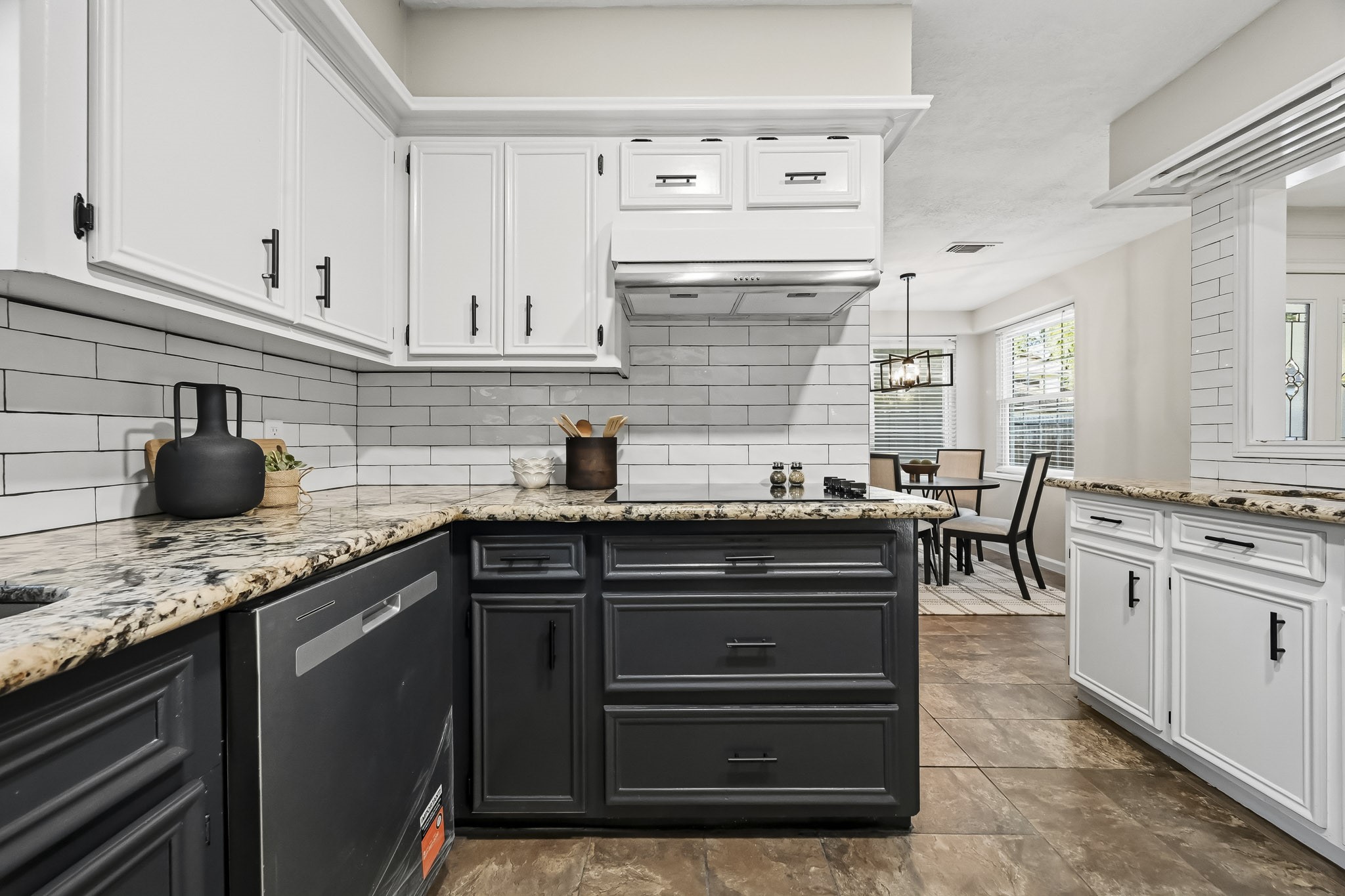 Stylish kitchen featuring a modern two-tone design with sleek black and white cabinetry, granite countertops, and a subway tile backsplash, seamlessly flowing into a bright dining area.