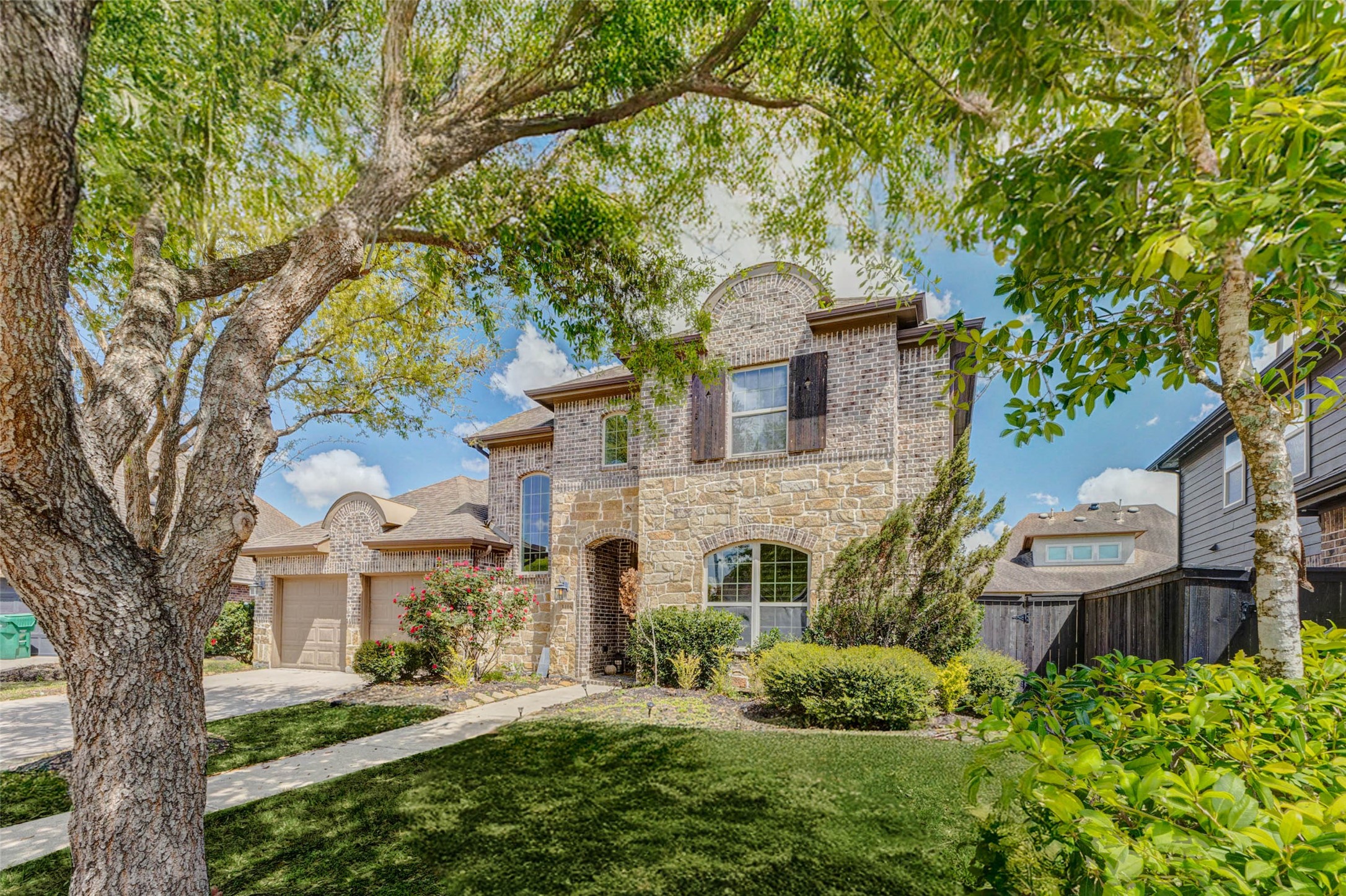 Framed by mature shade trees, this brick elevation immediately captures attention with its timeless architecture and inviting curb appeal. The arched entryway, detailed masonry, and charming shutters create a warm yet elegant first impression. Lush landscaping and a manicured lawn enhance the natural beauty of the setting, while the soft canopy of greenery provides both privacy and a welcoming sense of arrival to this beautifully maintained home.