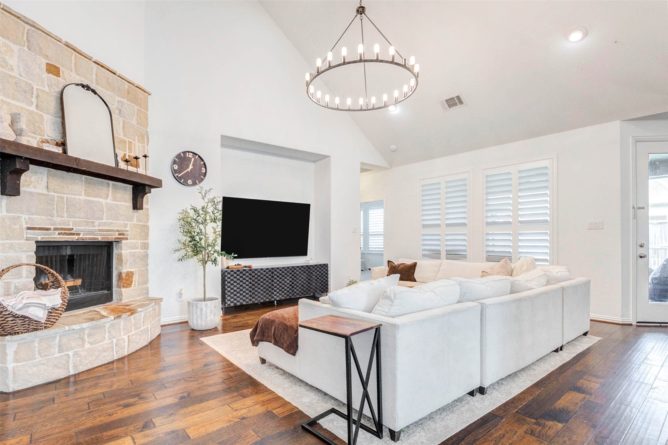 This expansive living room is filled with natural light and designed for both comfort and style. Soaring ceilings and a statement chandelier create an airy, open feel, while the stone fireplace adds warmth and character. Large windows with plantation shutters enhance privacy and elegance. The seamless flow into adjacent spaces makes this area perfect for entertaining or relaxing in a beautifully curated environment.
