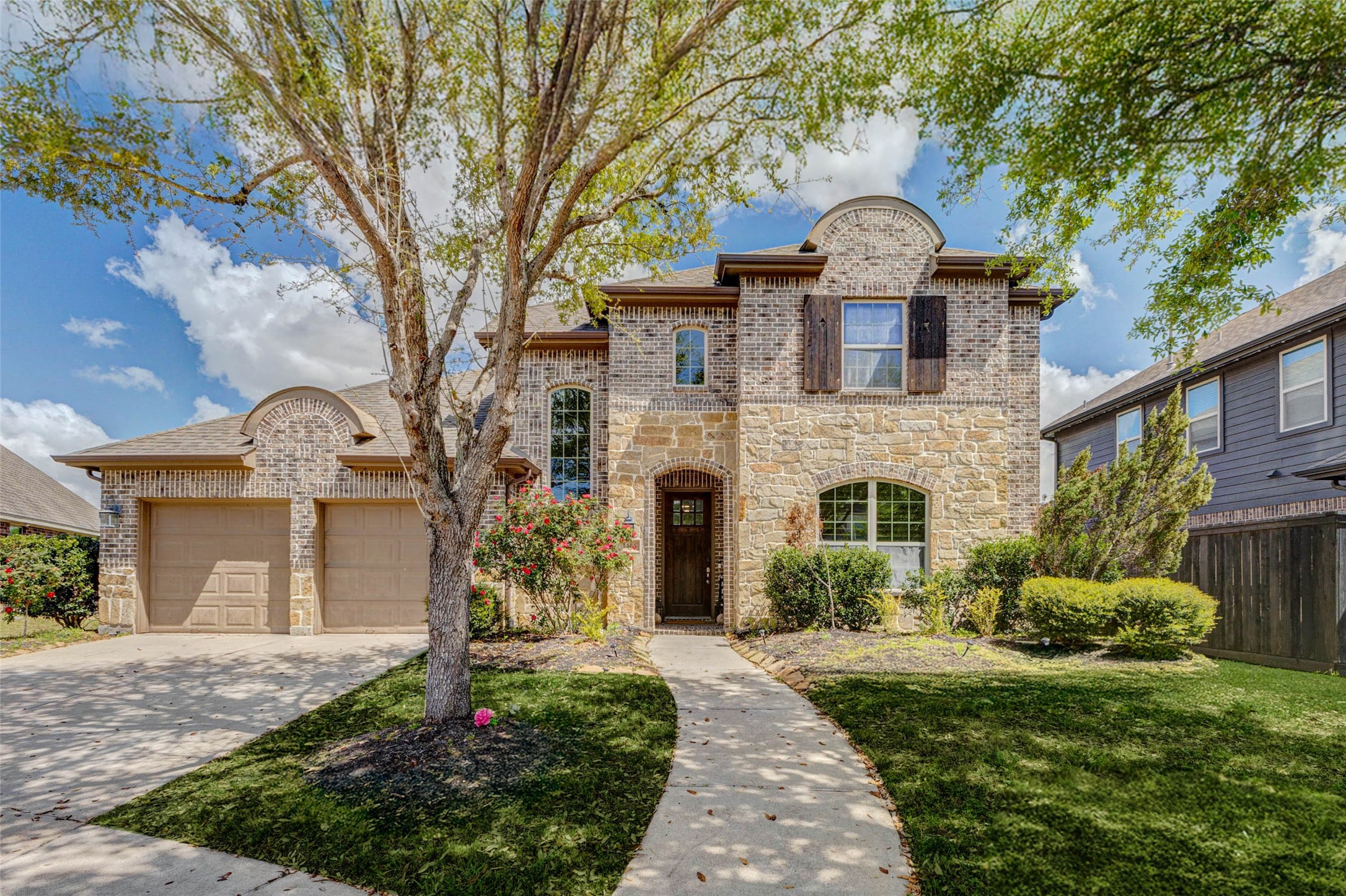 A perfectly balanced front elevation showcases classic design and thoughtful symmetry, highlighted by a grand arched entry. The winding walkway draws you toward the inviting front door, while blooming landscaping adds color and life. Large windows bring in natural light, and the combination of textures creates a sophisticated yet approachable exterior that sets the tone for the quality found throughout the home.
